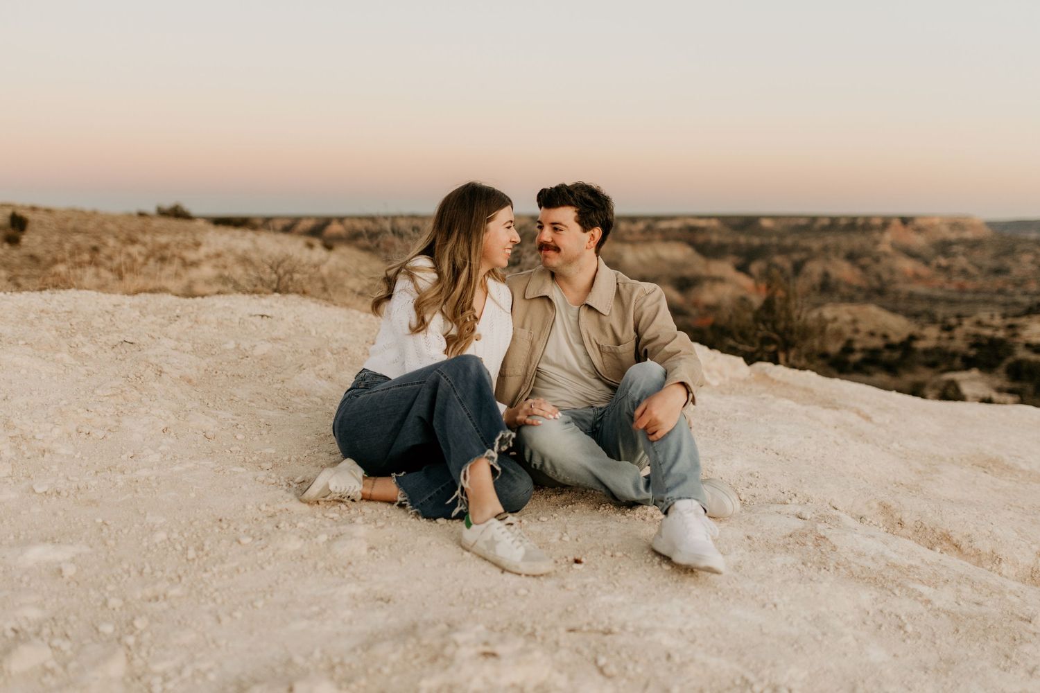 Couple sitting together on a rocky desert landscape at sunset, smiling and gazing at each other in casual outfits.