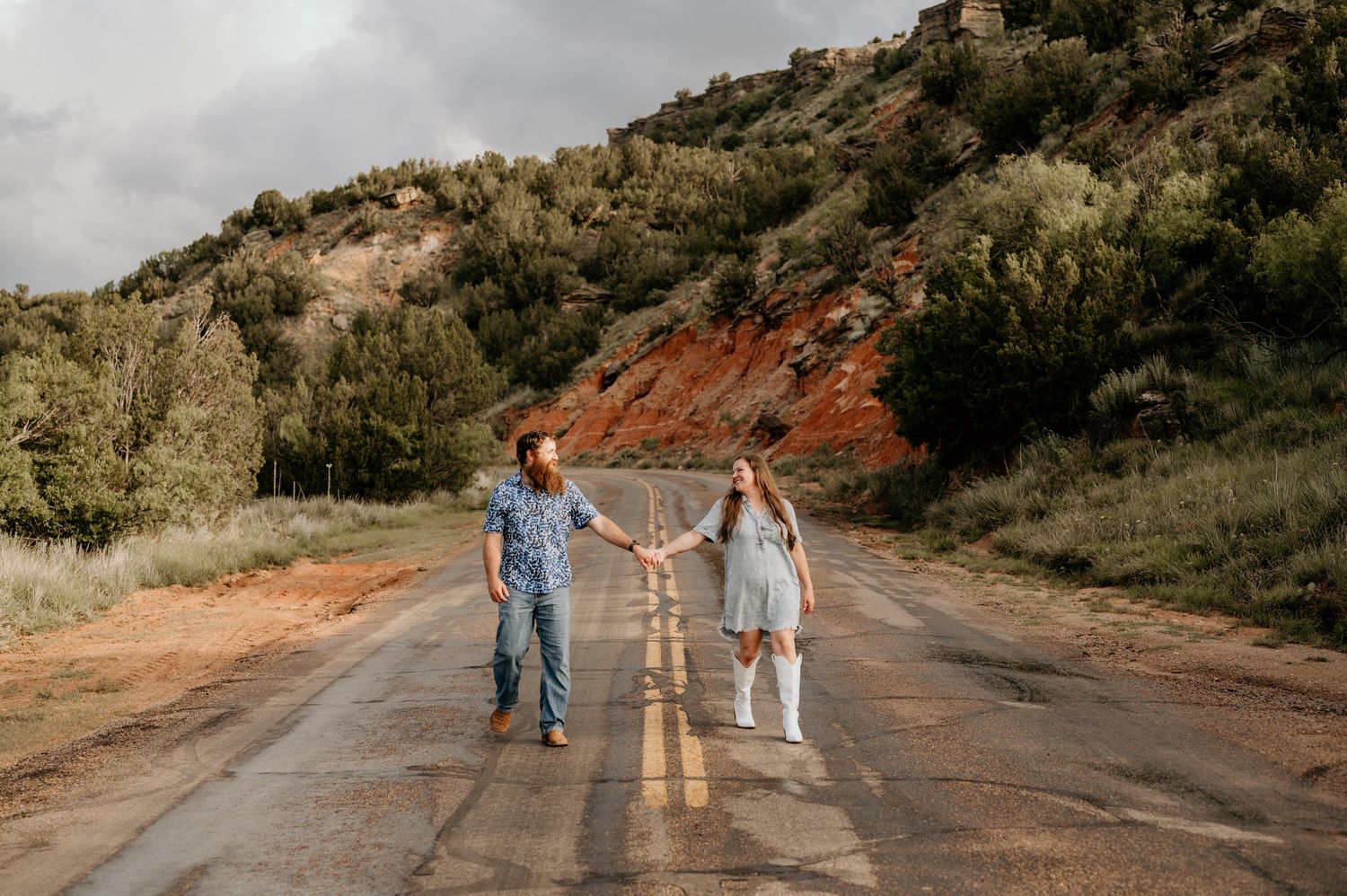 Couple holding hands on a scenic road surrounded by red rock hills and lush greenery under a cloudy sky.