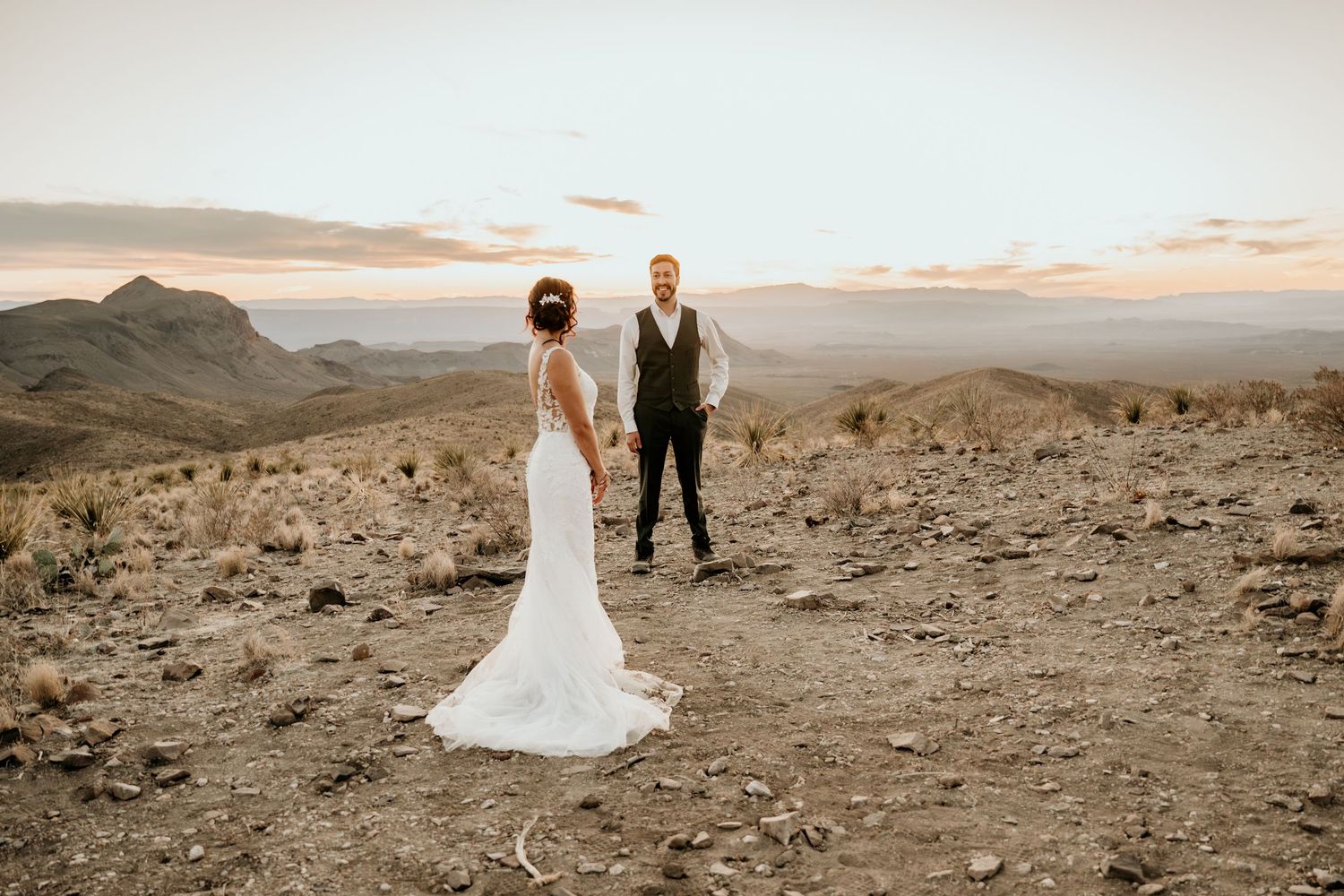 A couple in wedding attire poses against a dramatic desert mountain landscape at sunset.