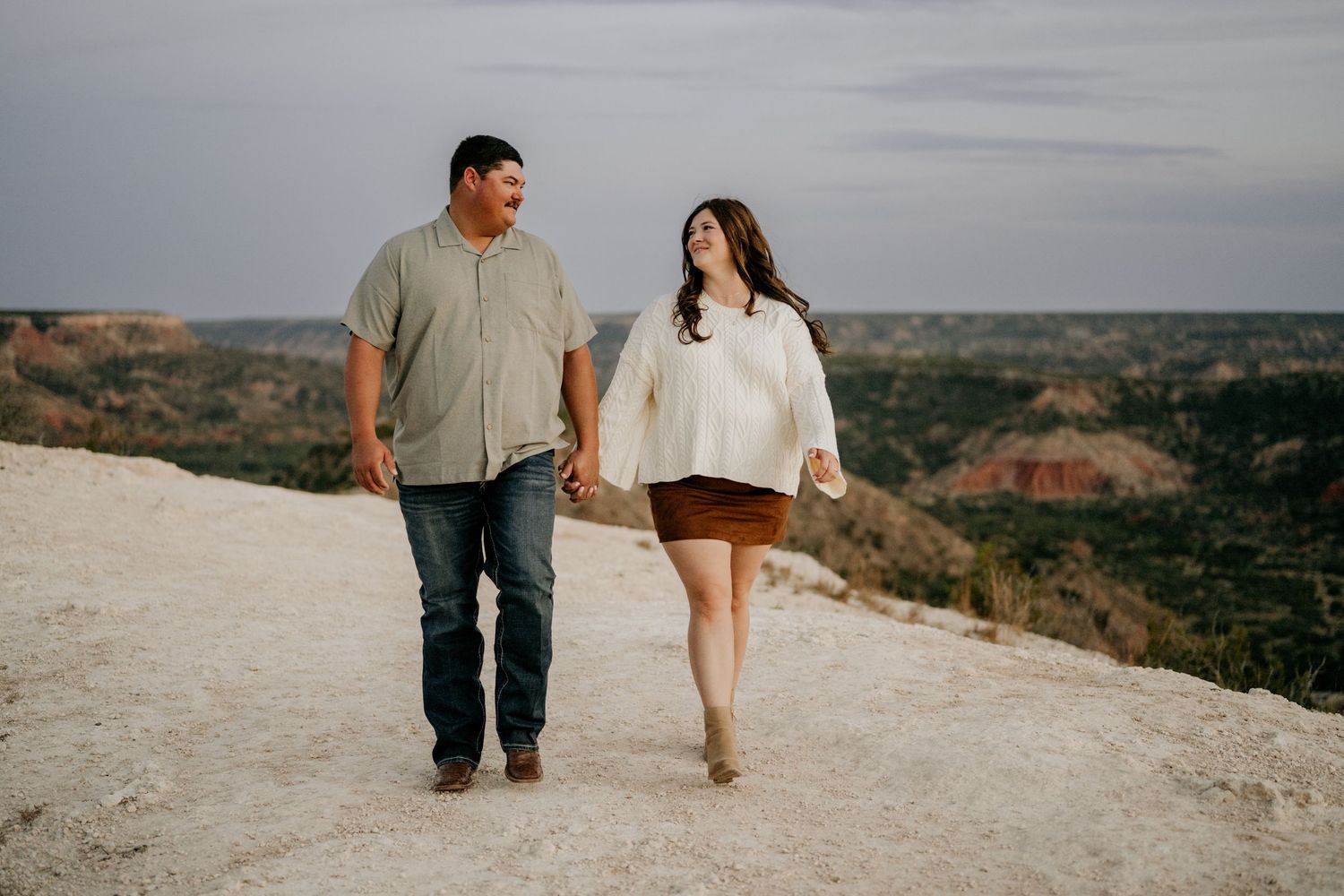 Couple holding hands walking on rocky overlook with canyon views in background.