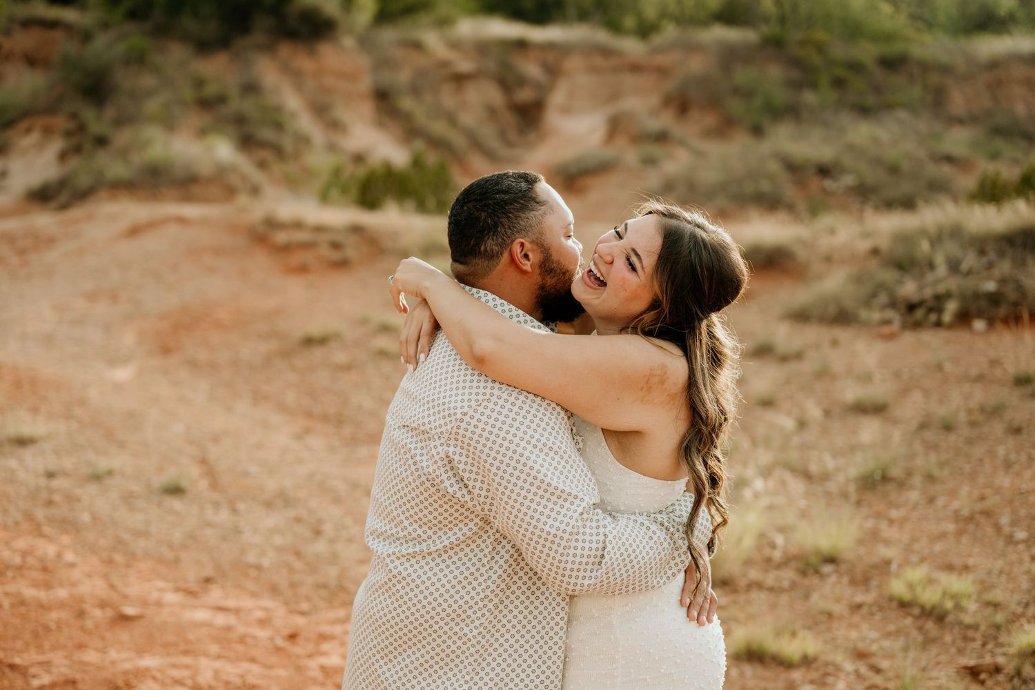 Couple embracing and kissing in a desert landscape with red dirt and rocky hills during an outdoor engagement photo session.