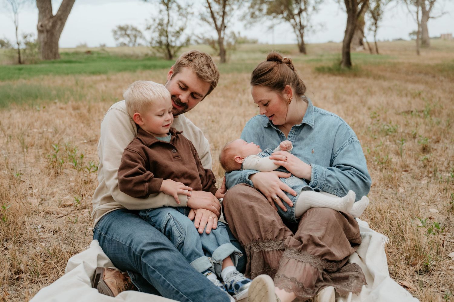 A smiling family of four sits together on a blanket in a dry grassy field, with a toddler and a newborn baby in their parents' arms.