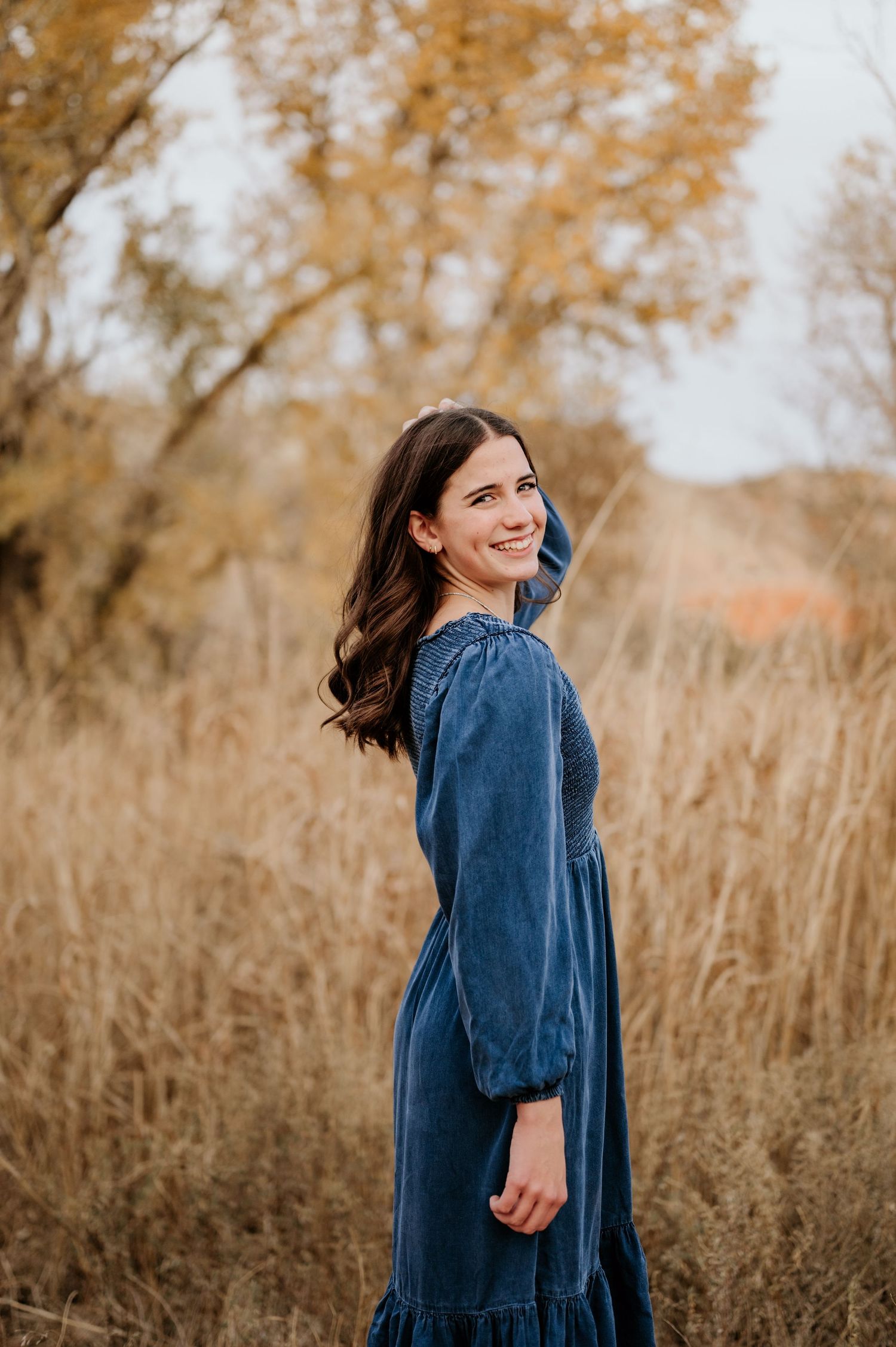 Young woman in blue denim dress smiling over shoulder in autumn field with golden grass and trees.