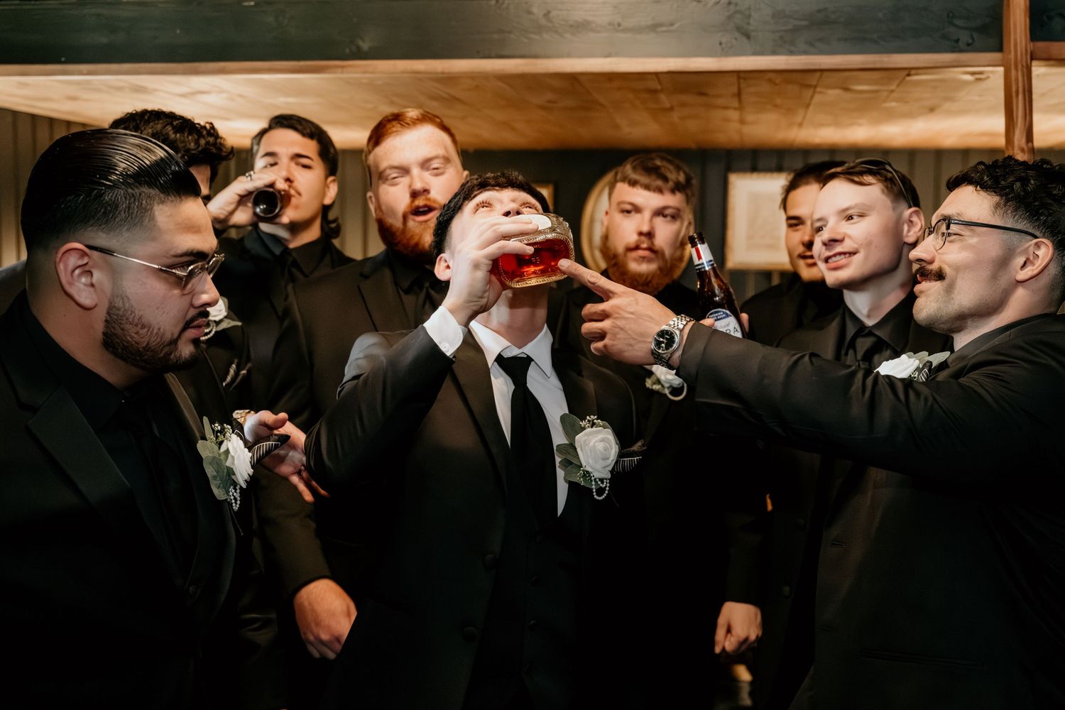 Groomsmen in black suits share laughter and candid moments at an indoor wedding venue.