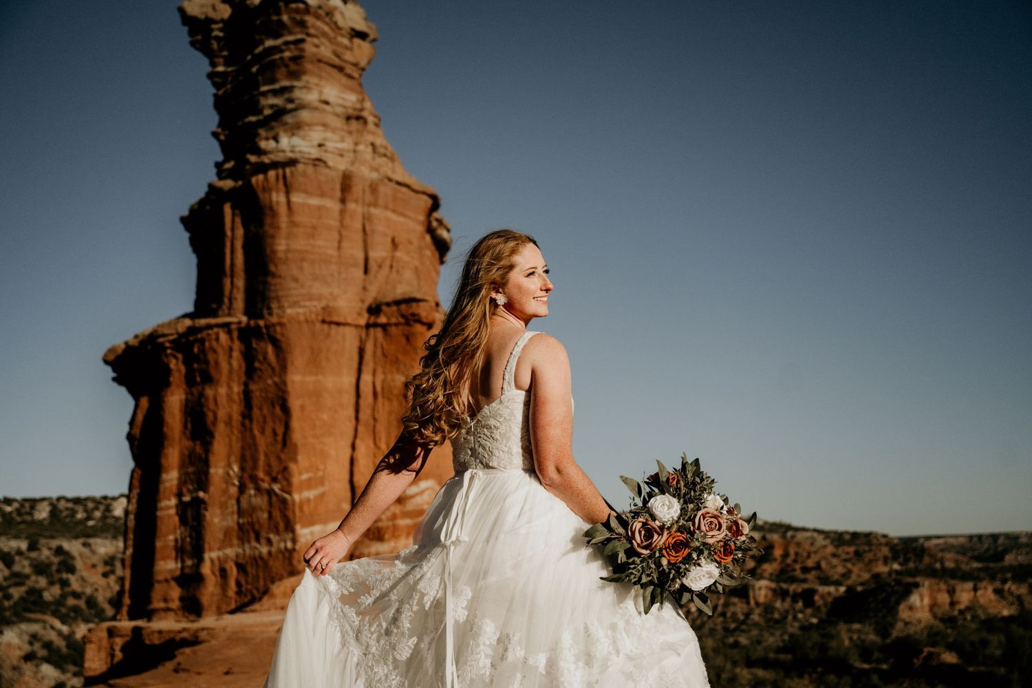Bride in lace wedding gown holds bouquet beside red rock formation at golden hour in canyon landscape.