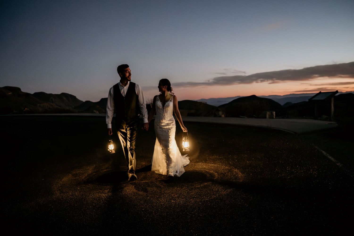 Silhouetted figures hold lanterns against a dramatic sunset in the desert mountains.