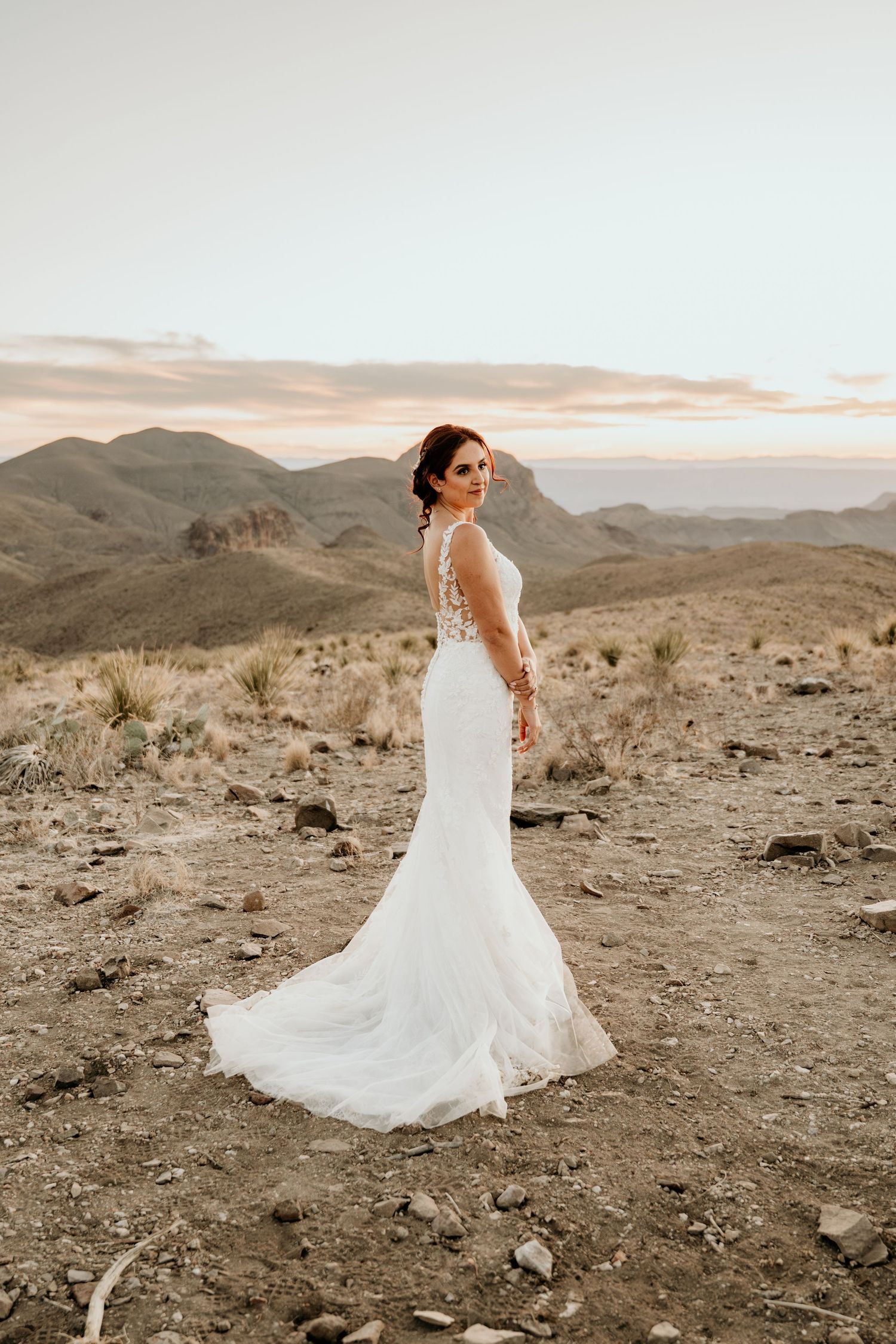 A bride in a flowing white dress twirls on rocky desert terrain at golden hour.
