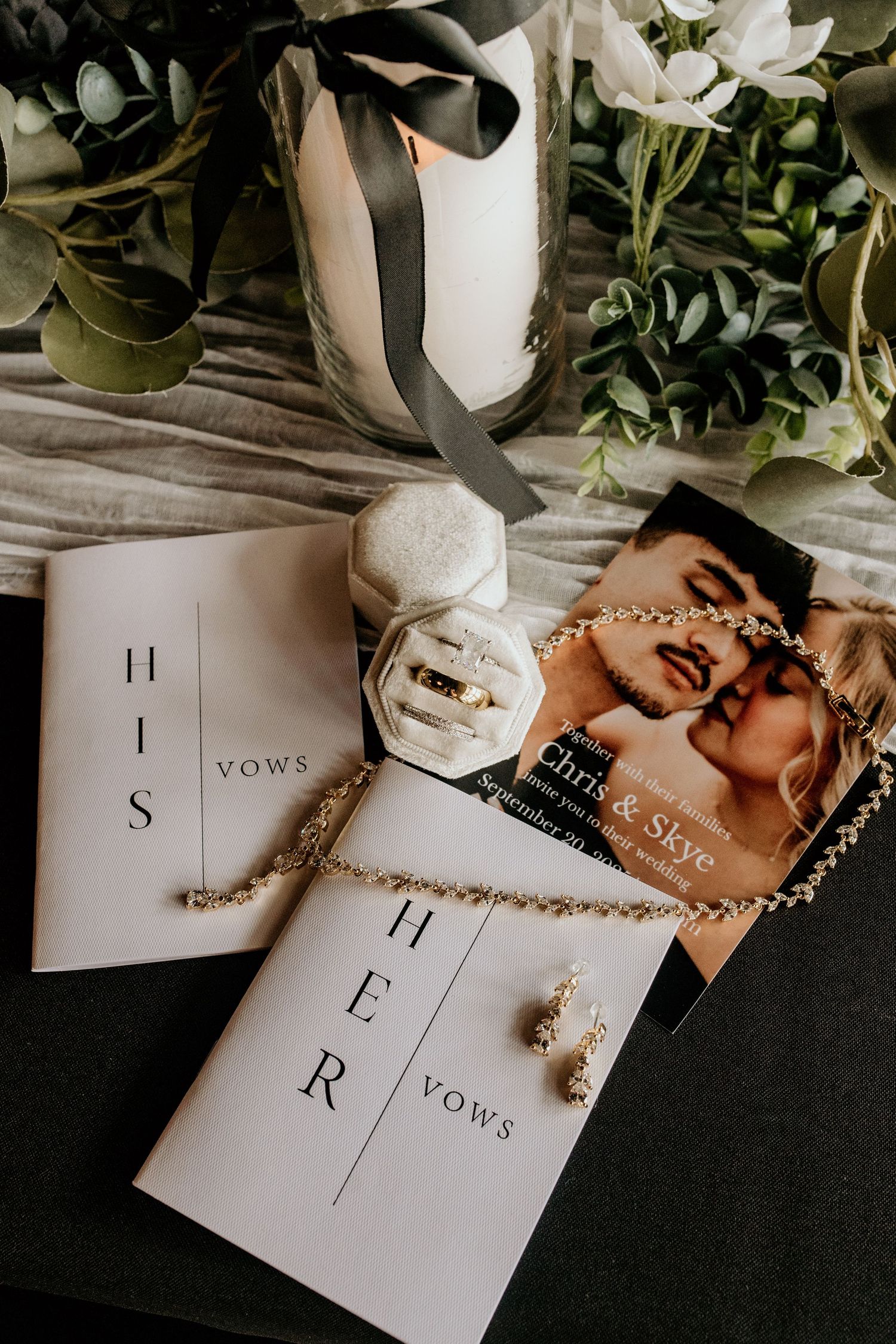 Wedding vow books and jewelry arranged with greenery and black ribbon for detail photography.