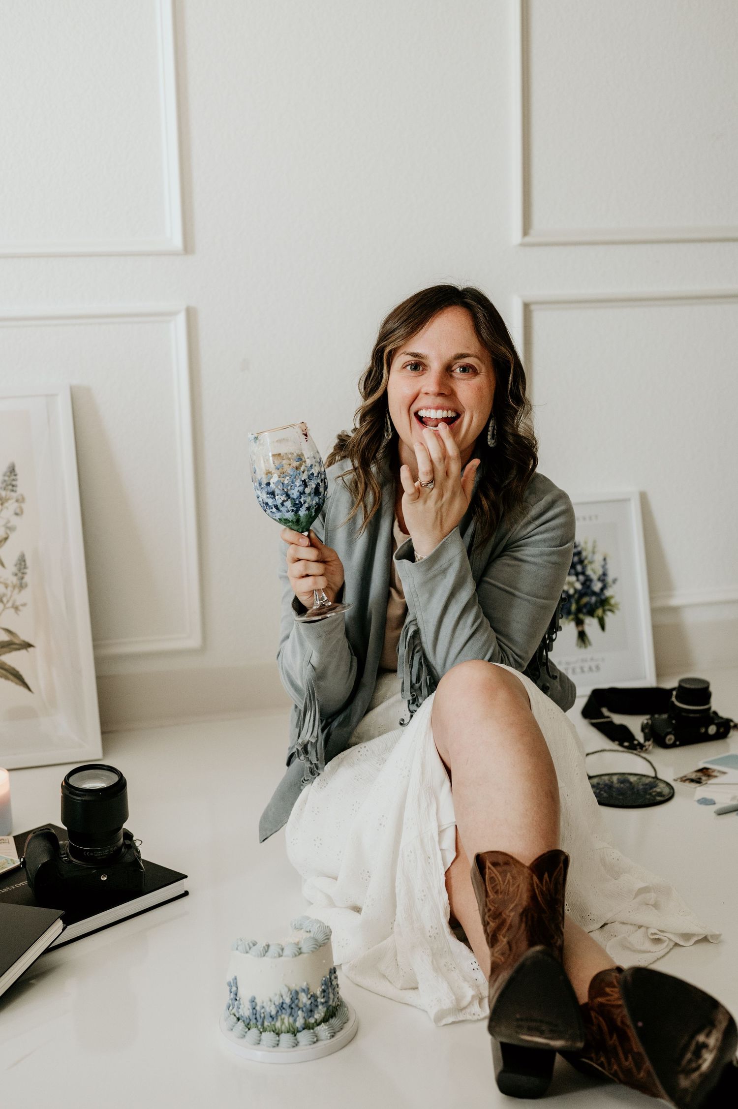 Woman laughing while holding a floral wine glass, sitting beside a small cake in a bright white studio setting.