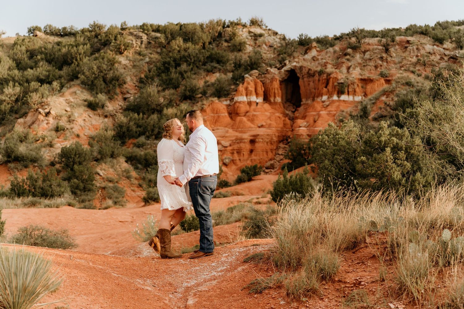 Couple embracing on red dirt trail at Palo Duro Canyon, surrounded by vibrant rust-colored cliffs and desert shrubs.
