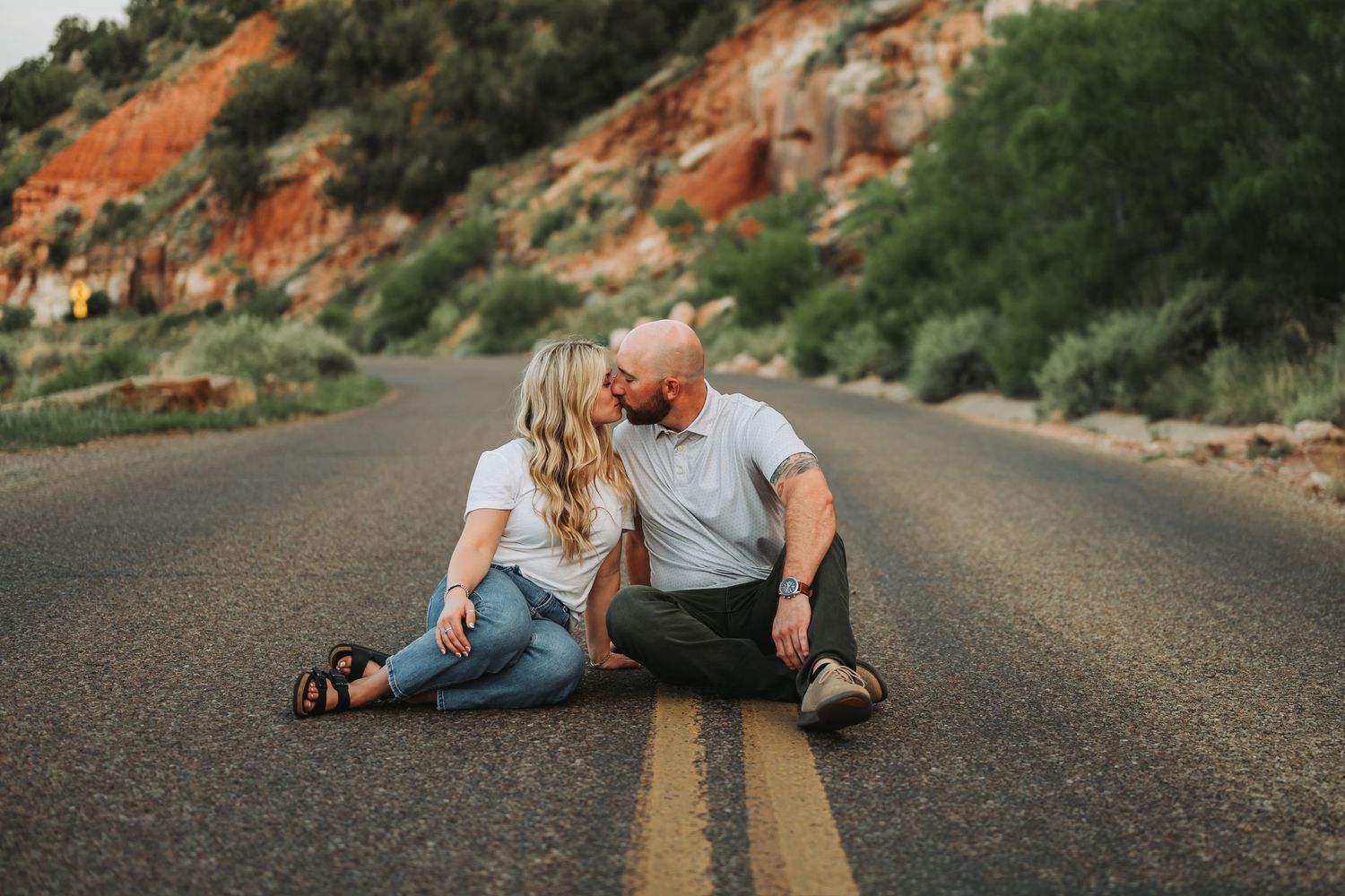 Couple sitting on a road kissing, surrounded by red rock canyon landscape during golden hour.