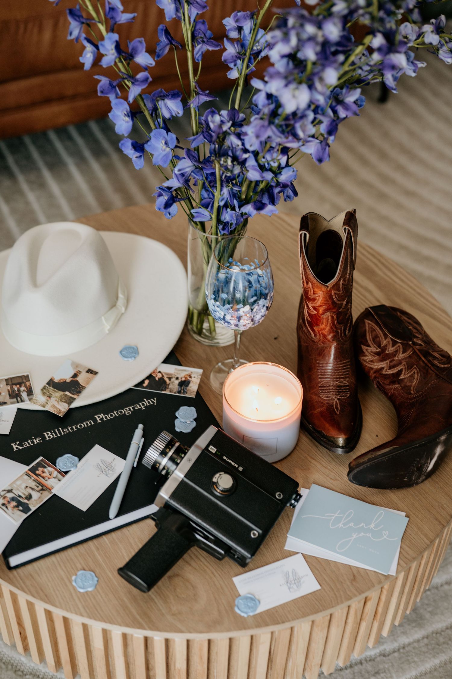 Wedding detail flatlay with blue flowers, cowboy boots, and photo albums on wooden surface.