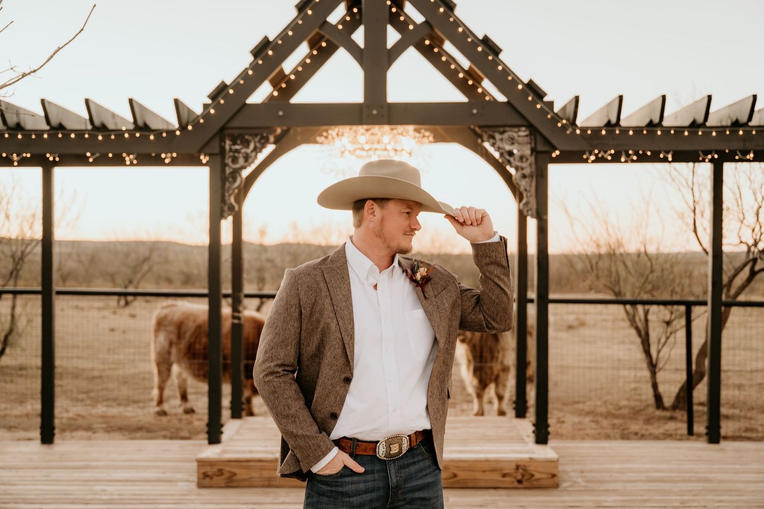 A person in western attire stands under a rustic wooden pergola at sunset.