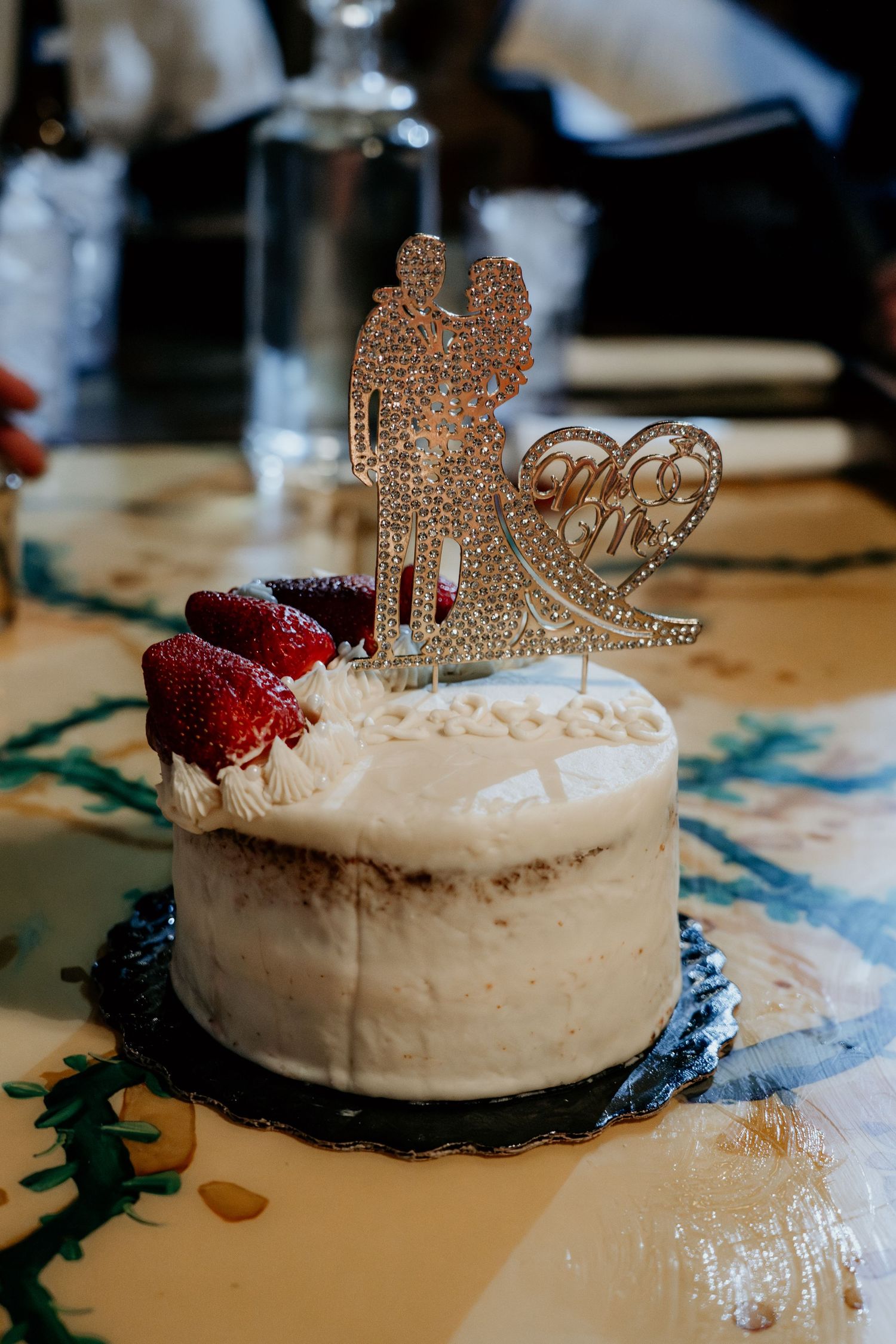 White wedding cake decorated with a sparkly topper and red berries on a patterned tablecloth.