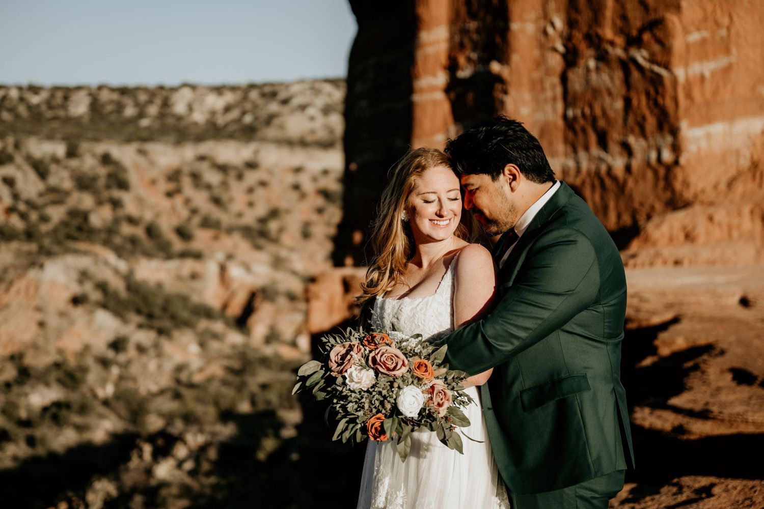 Bride and groom embrace at red rock canyon overlook, bride holding boho floral bouquet in white gown, groom in green suit.