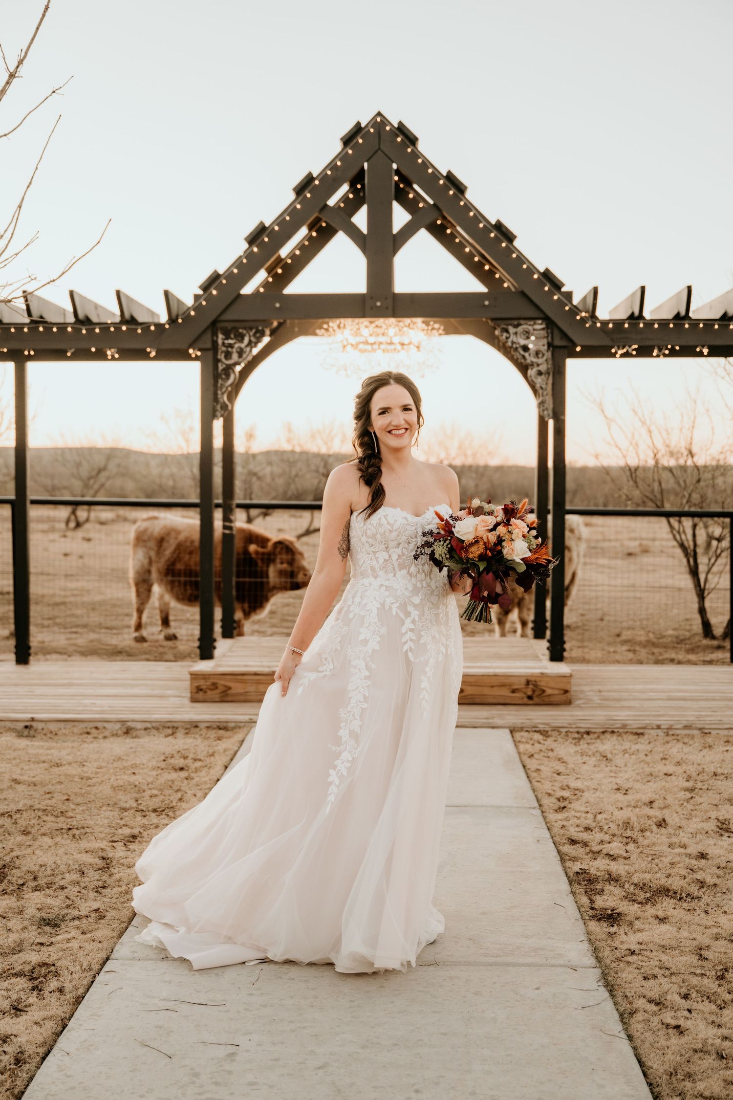 A bride in a flowing lace gown poses under a wooden pergola decorated with string lights.