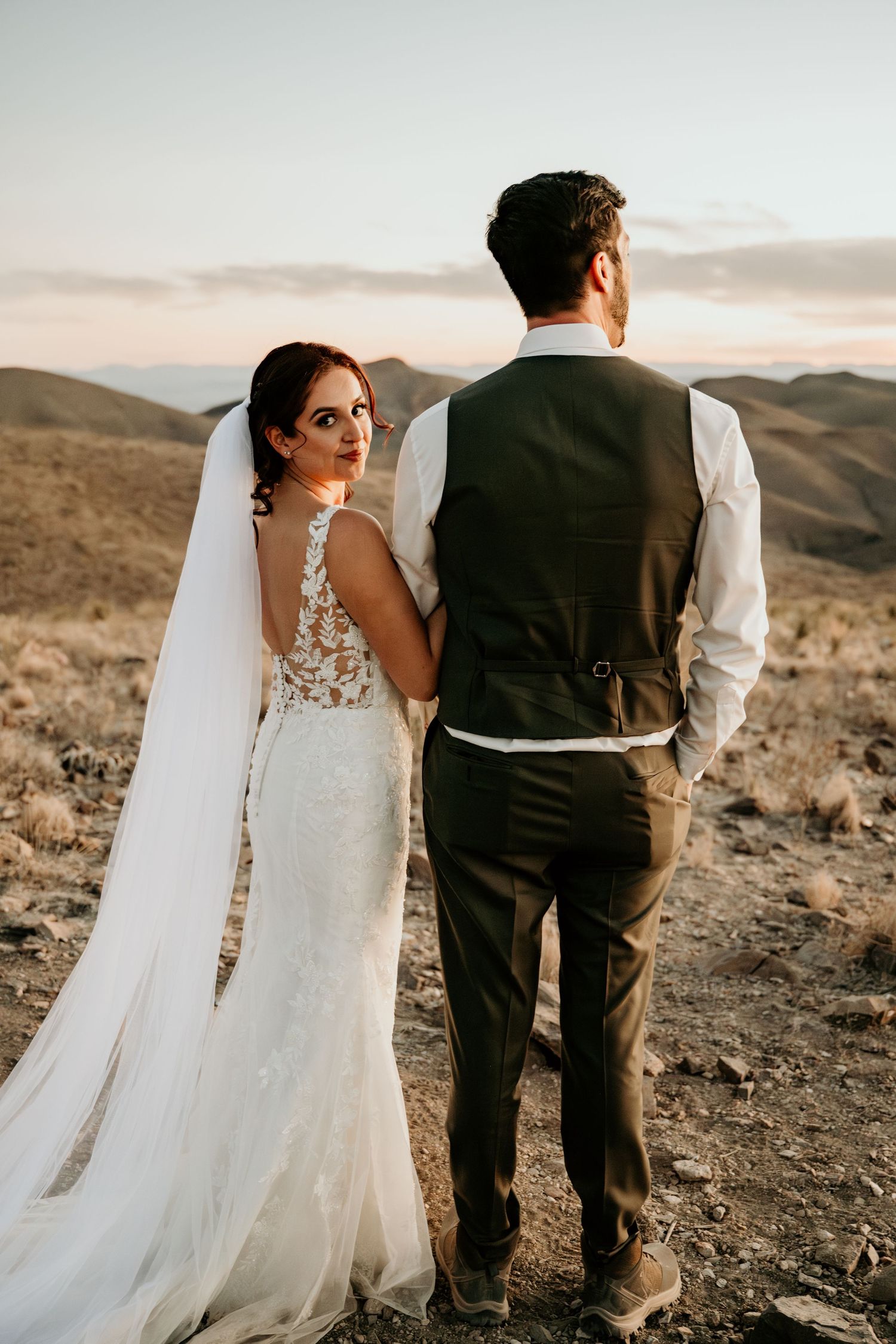 Wedding couple standing together on rocky desert terrain at sunset.