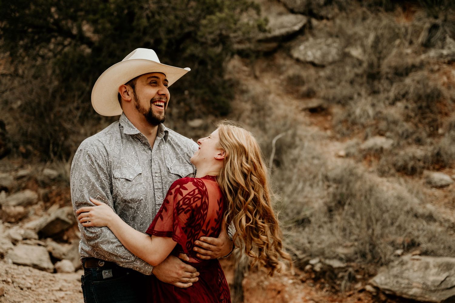 Couple in rustic outdoor setting, man in cowboy hat laughing as woman in red lace dress embraces him among desert rocks.