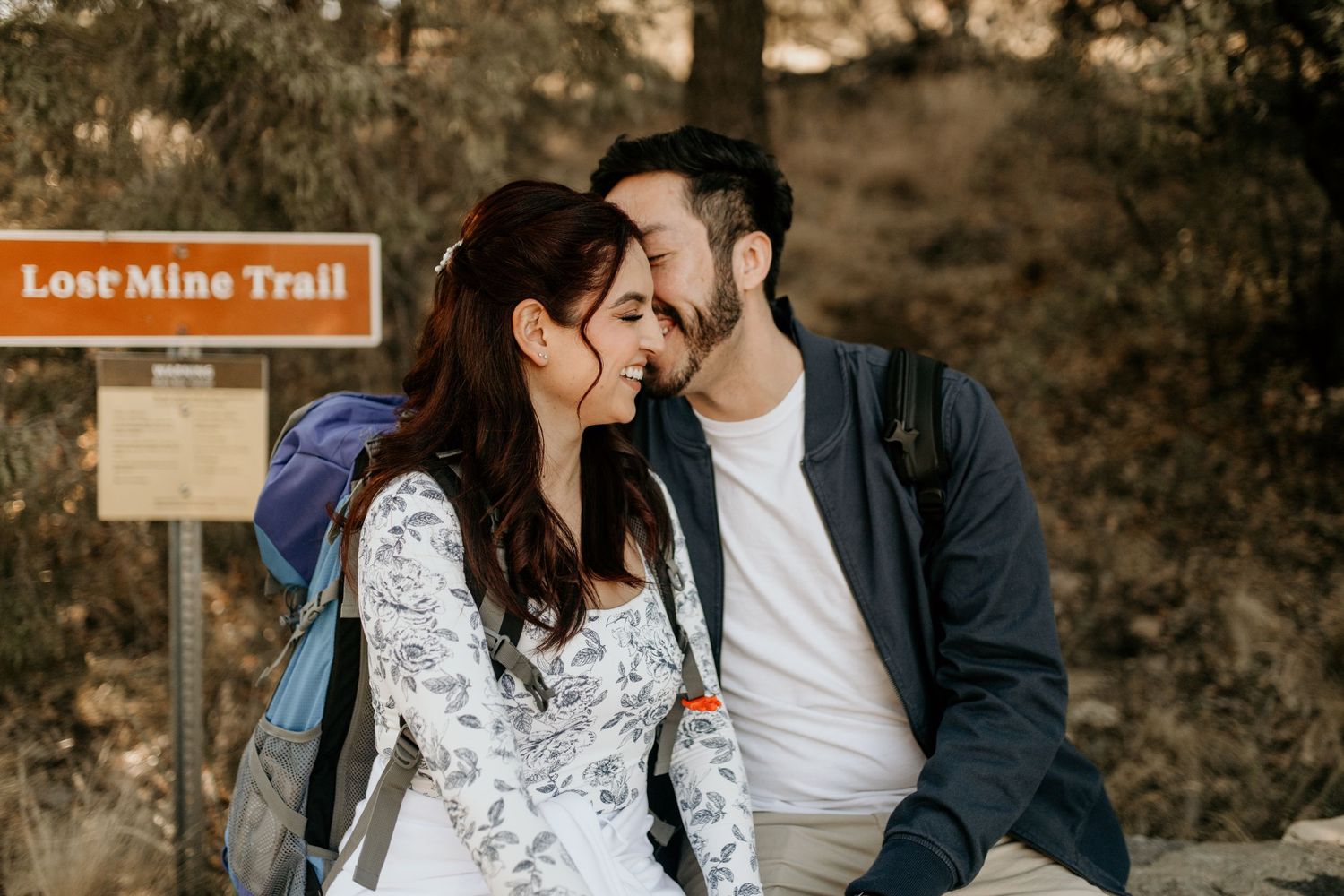 Couple sharing a romantic moment on the Lost Mine Trail during golden hour, with a wooden trail sign visible.