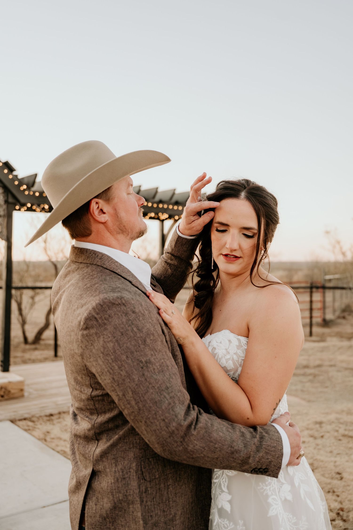 A couple in western wedding attire share an intimate moment under string lights at dusk.