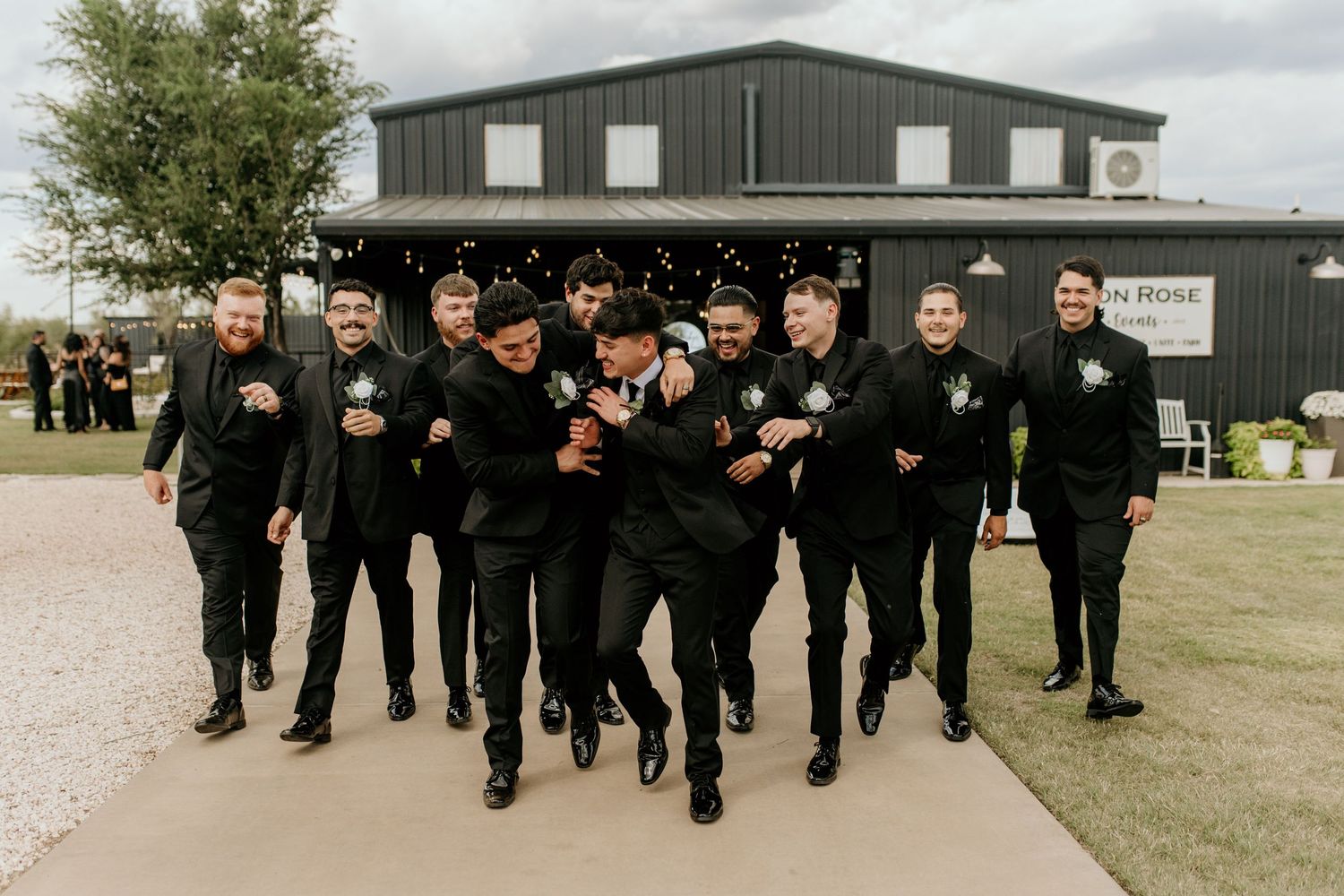 Wedding party in black suits poses playfully outside a modern industrial building.