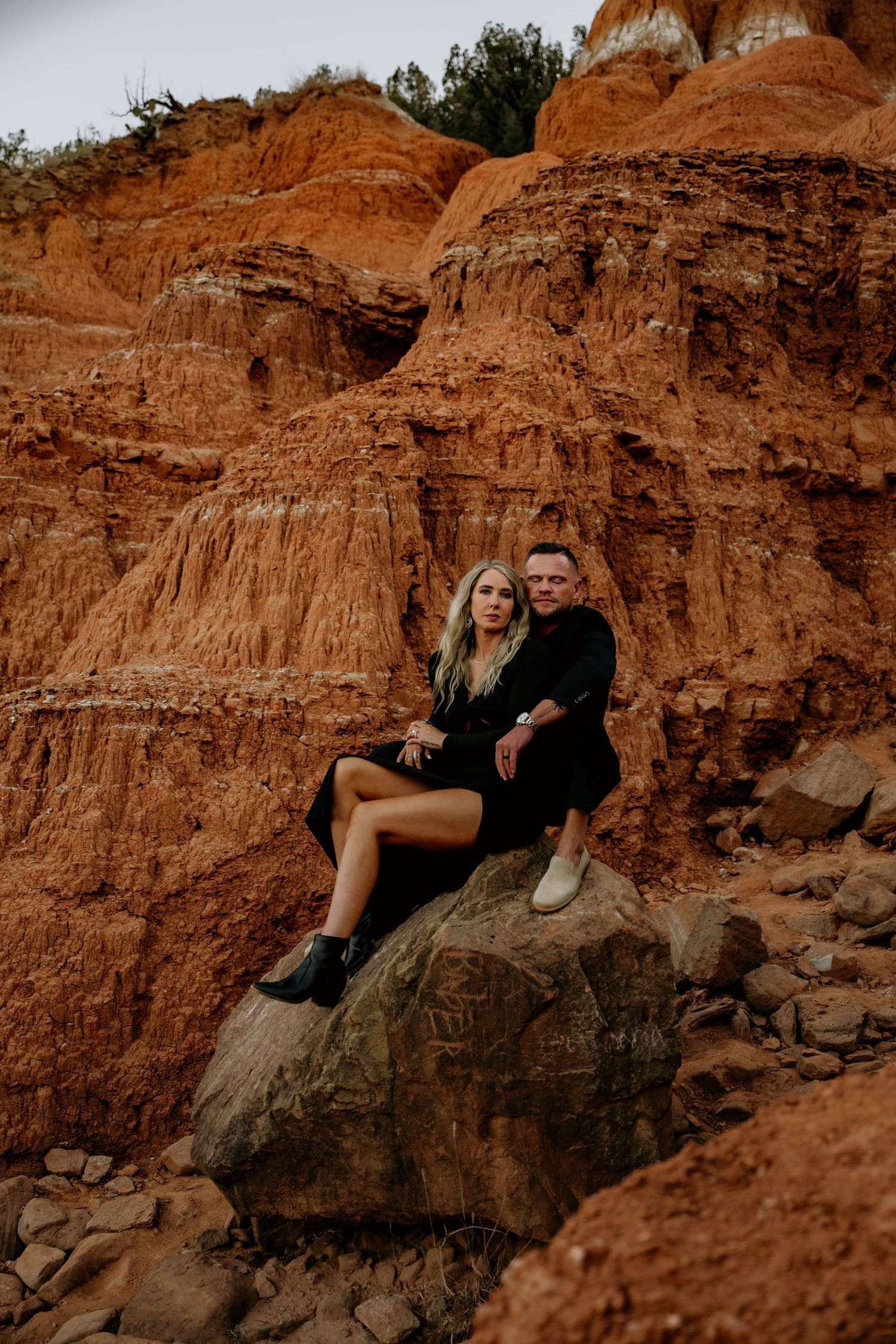 Couple in black outfits sitting on a rock against dramatic red canyon formations for an engagement photo session.