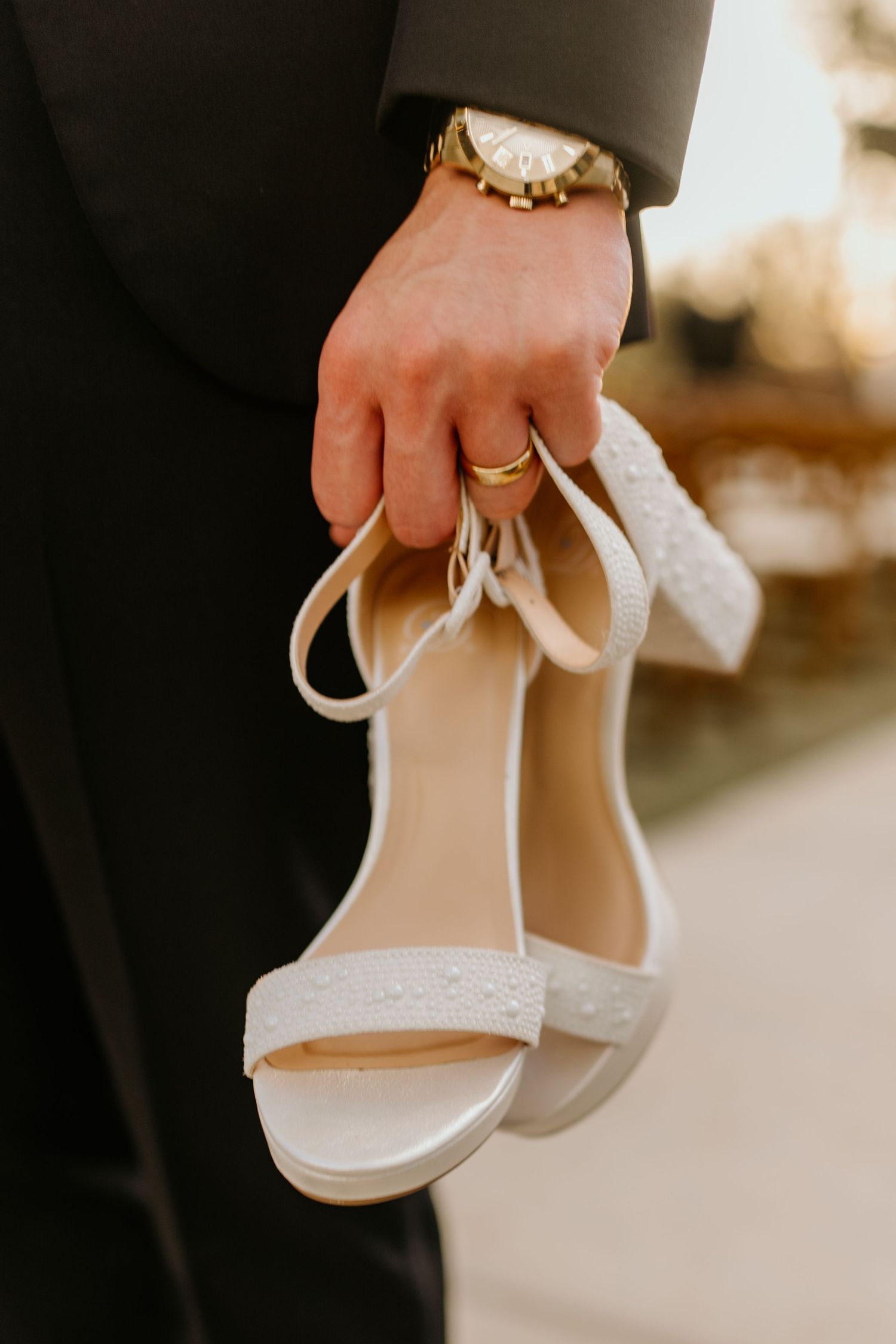 A hand wearing a wedding ring holds elegant white bridal sandals.