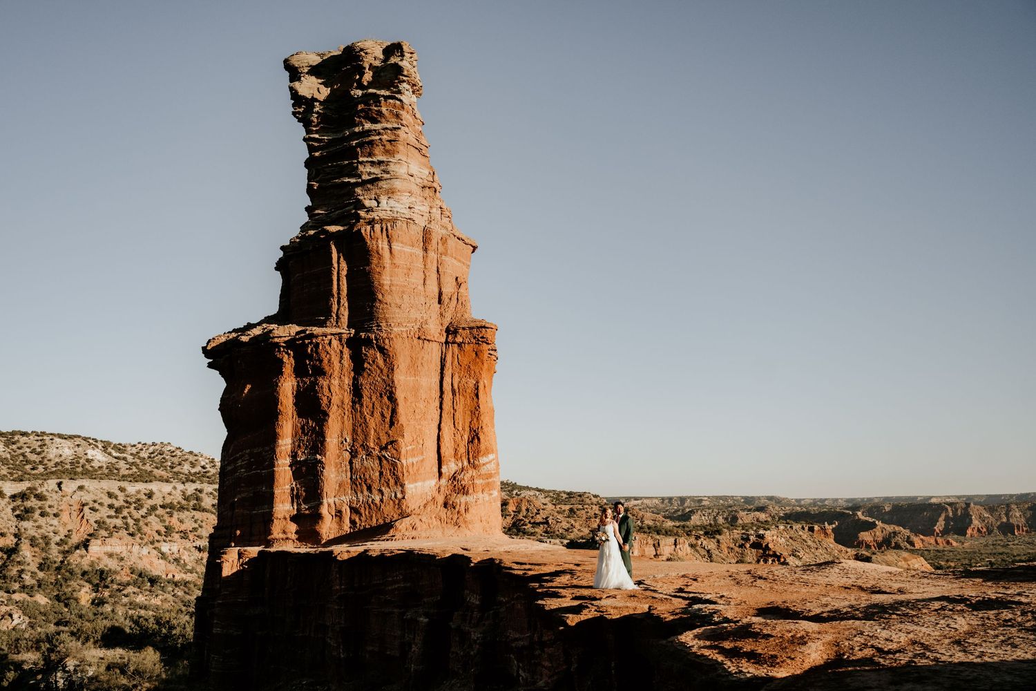 Couple in wedding attire standing beside a towering red rock formation in a vast desert landscape under a clear blue sky.