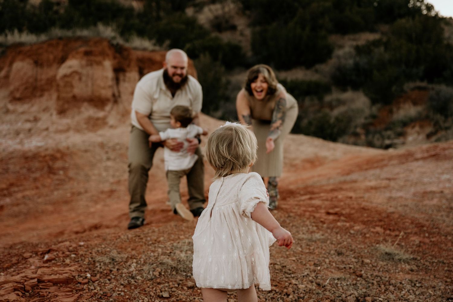 Family with two young children laughing and playing together on a red dirt trail in a desert canyon landscape.