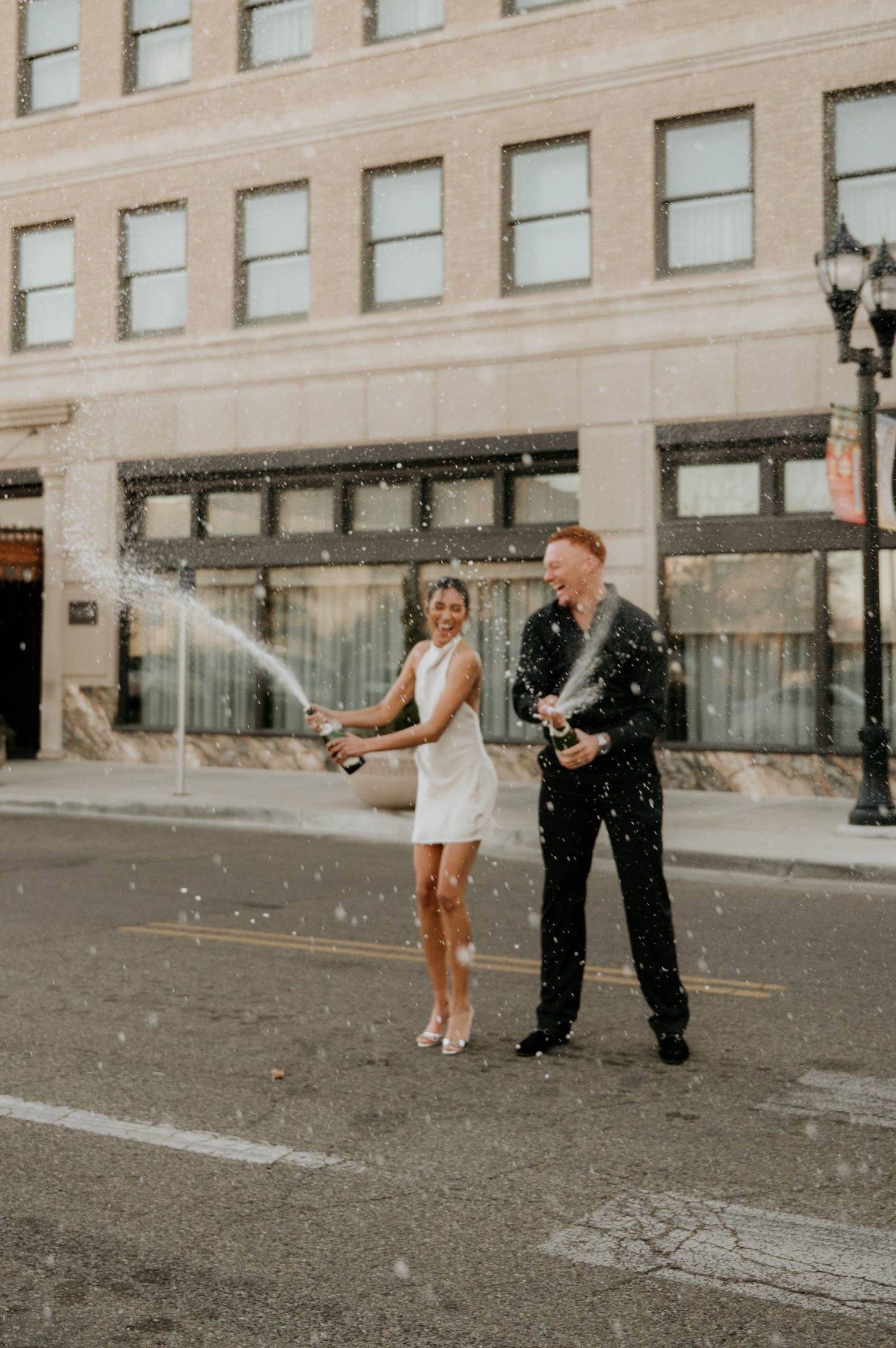 A couple celebrates with champagne on a city street in front of a vintage building.