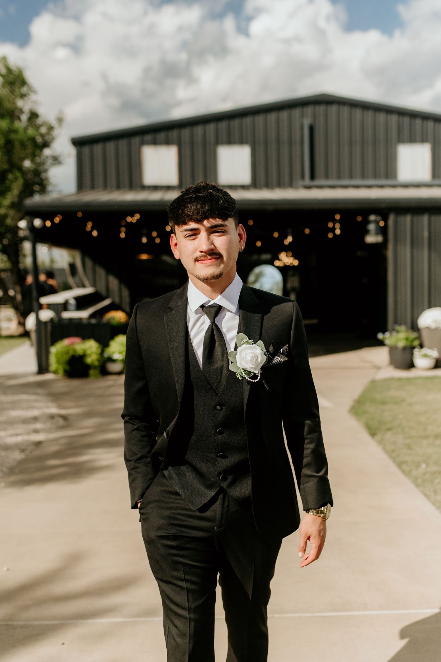 Groomsmen in formal black tuxedos walk along a concrete path near a metal building.