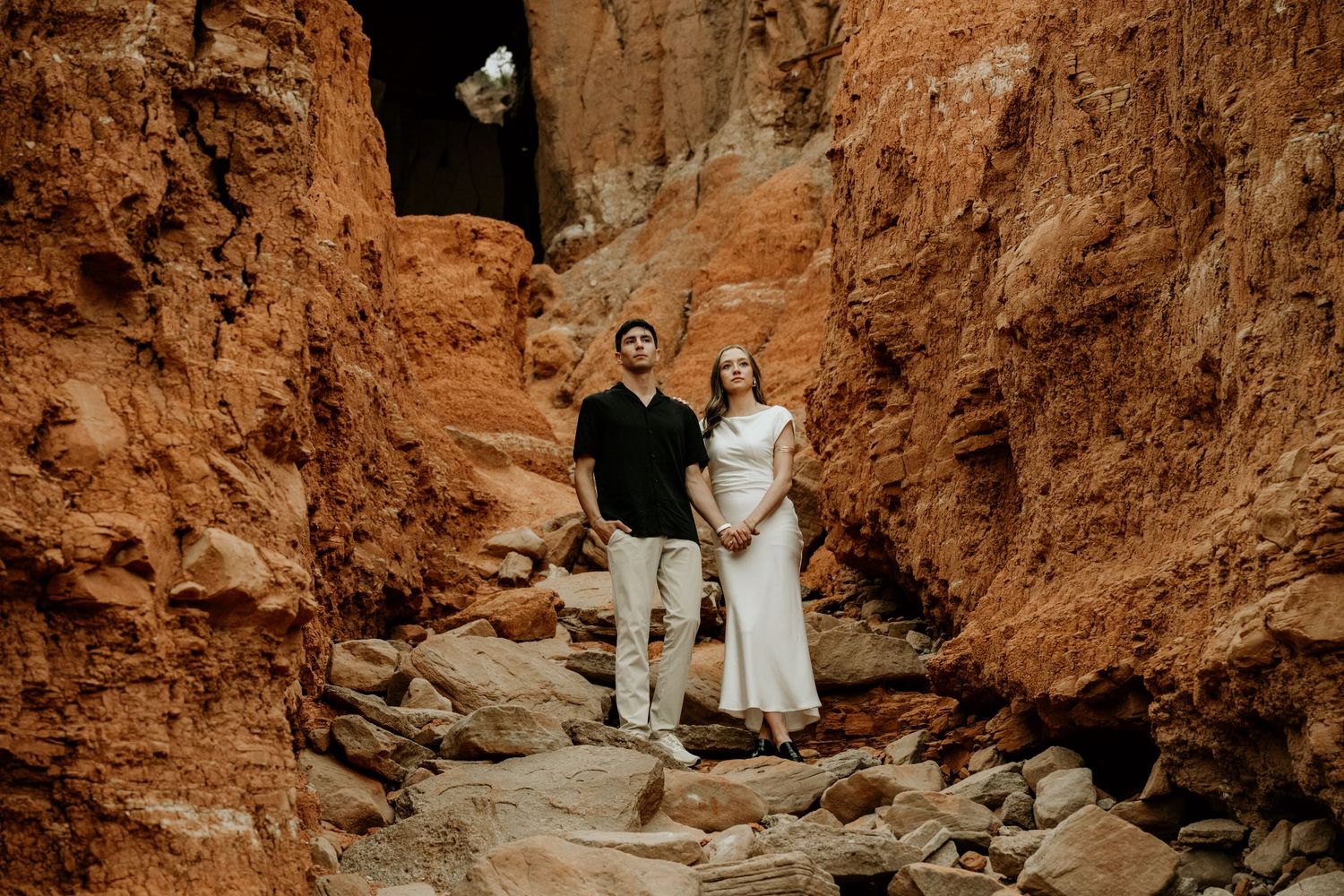 Couple standing between red rock canyon walls, man in black shirt and woman in white dress, outdoor engagement photo.