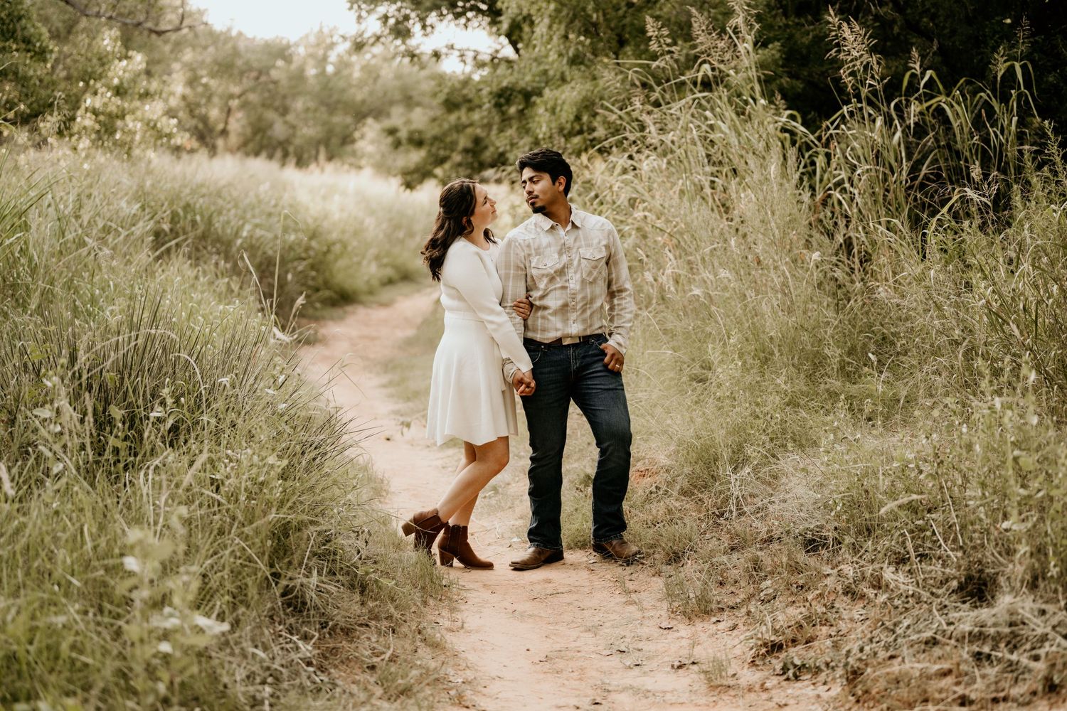 Couple holding hands on a rustic dirt path surrounded by tall green grass in a romantic outdoor engagement photo.
