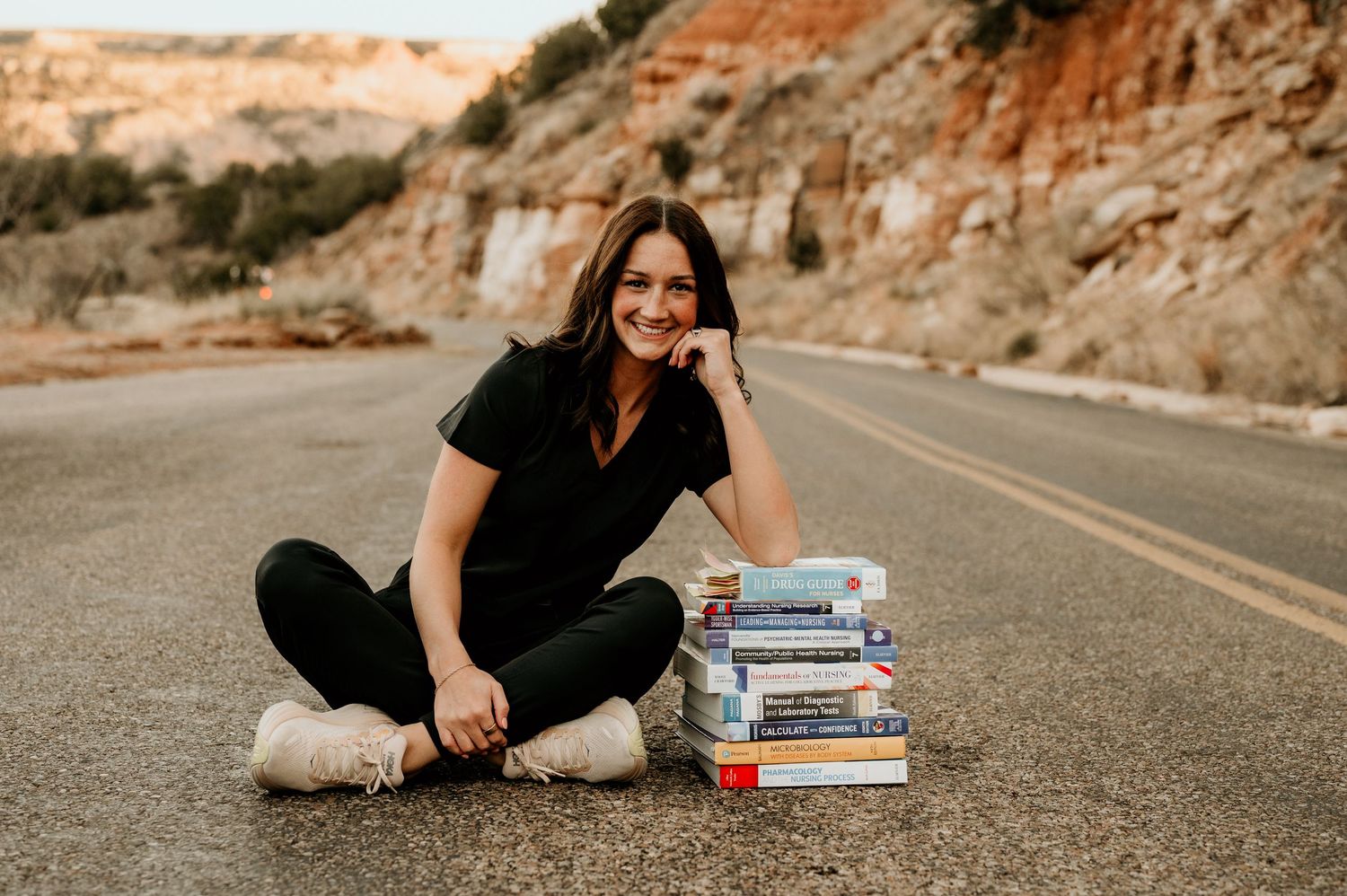 Young woman sitting cross-legged on a road next to a stack of books, smiling, with scenic rocky hills in background.