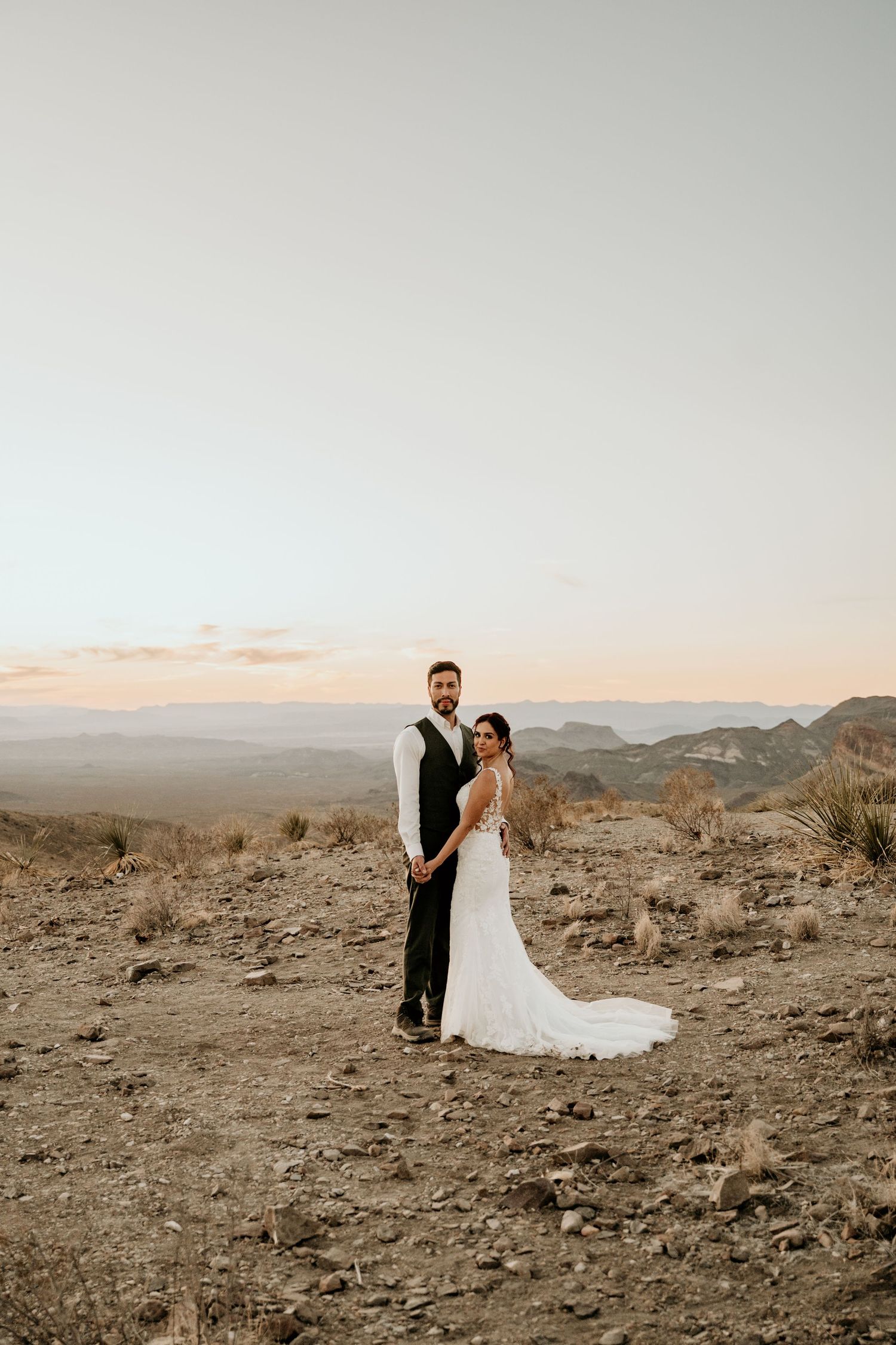 Wedding couple stands together on rugged desert terrain with mountains in the distance.