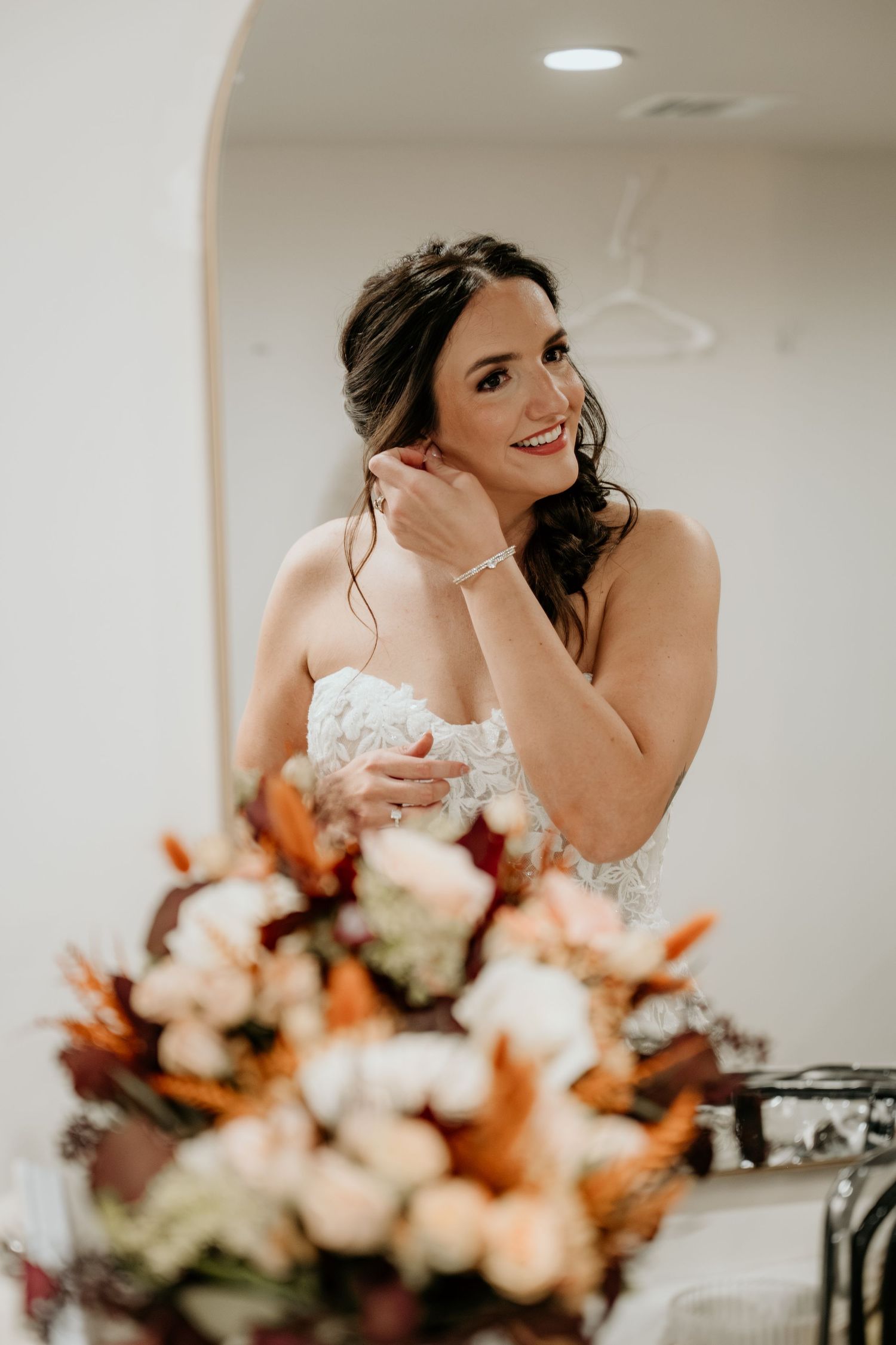 A bride in a white lace gown adjusts her jewelry while standing next to an autumn-colored floral arrangement.
