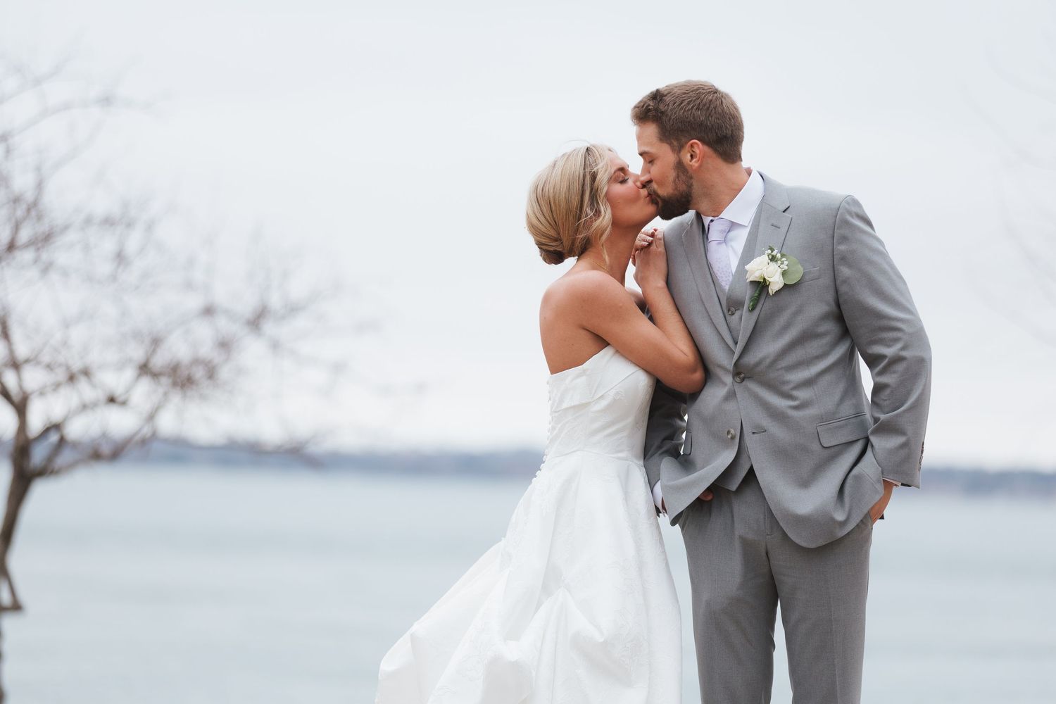 Bride and groom share a romantic kiss outdoors near waterfront, bride in white gown, groom in gray suit.