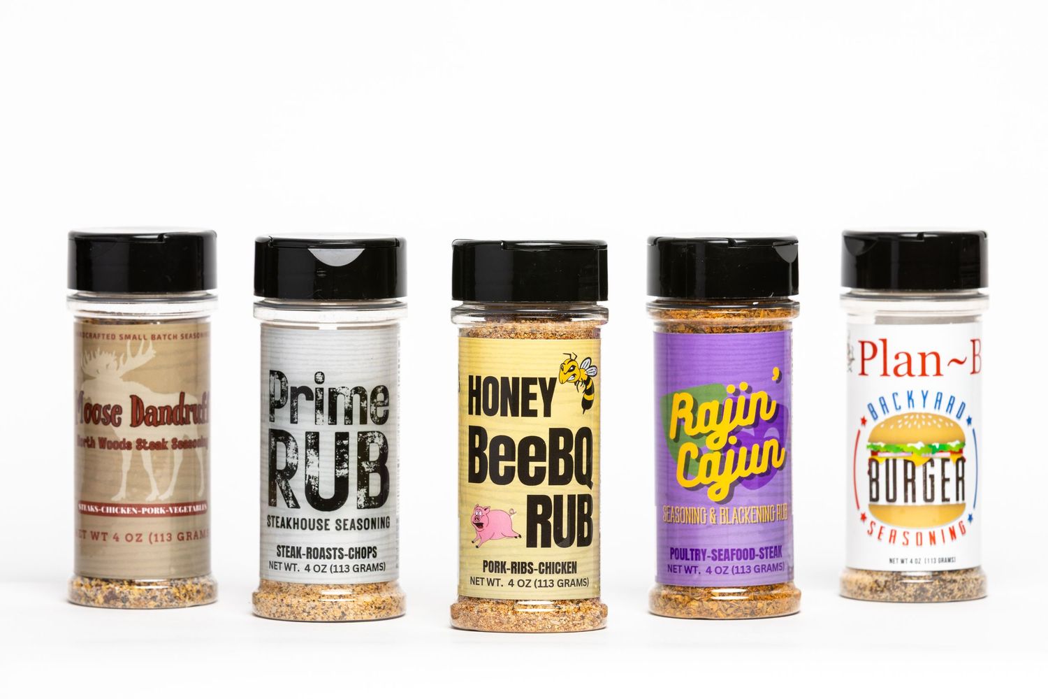 Five assorted BBQ spice rub seasoning bottles including Honey BeeBQ Rub and Rajin Cajun on white background.