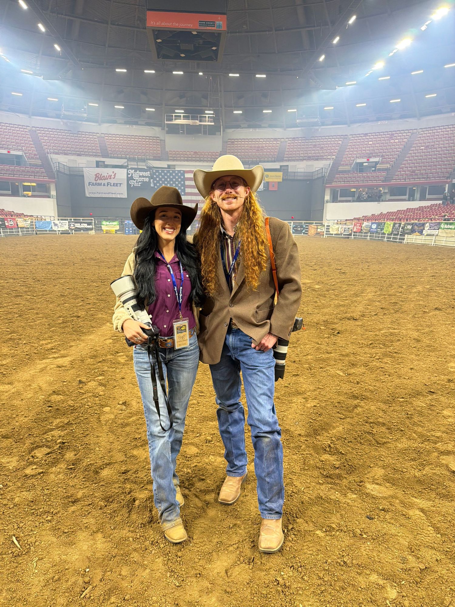 Two people in western attire and cowboy hats pose in a rodeo arena with stadium seating in the background.