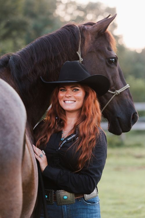 Smiling woman with red hair in black cowboy hat and shirt stands beside a dark brown horse outdoors.