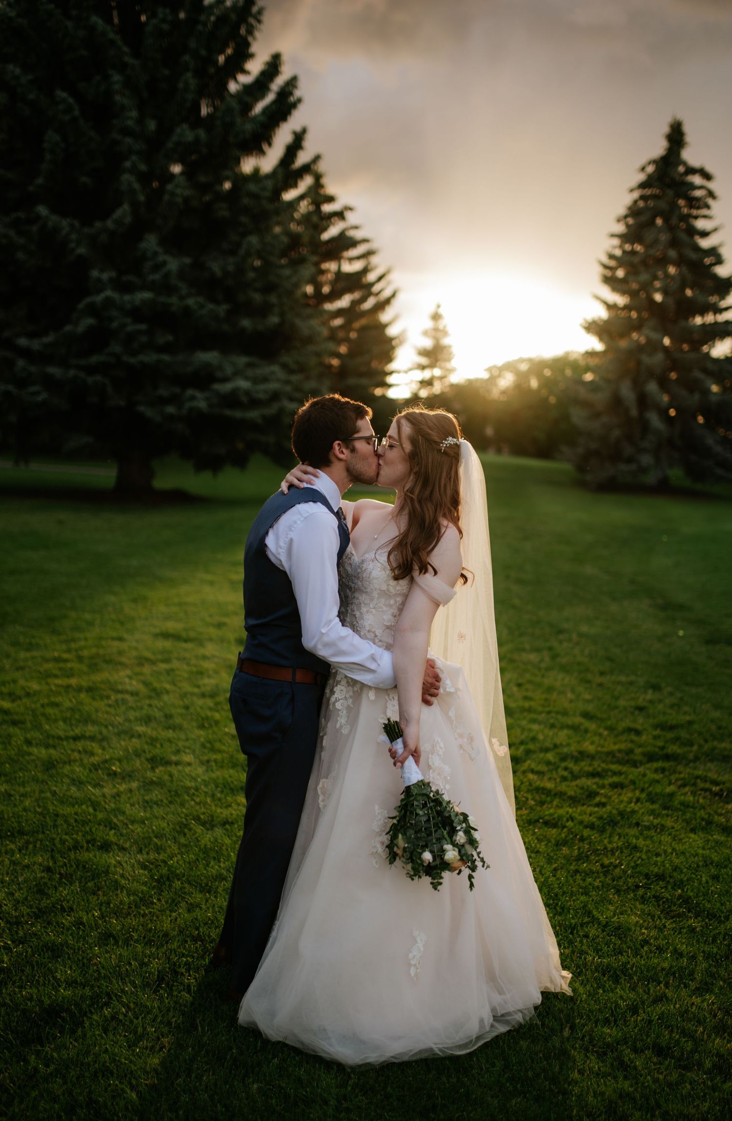 Couple in formal wedding attire share romantic moments against sunset and evergreen trees in a lush green field.