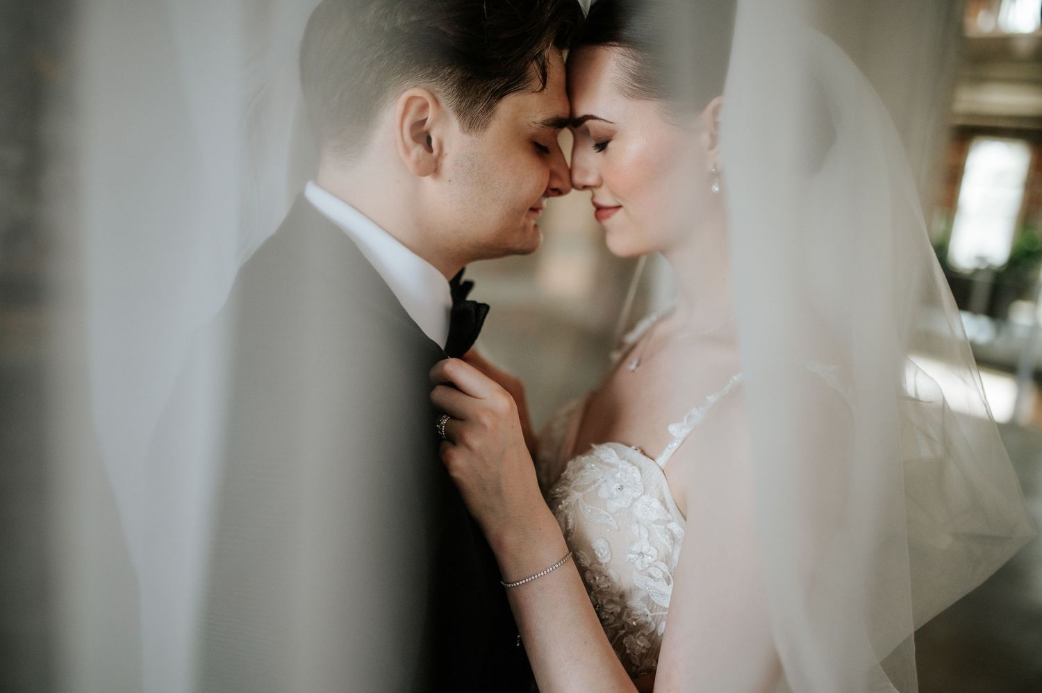 Intimate wedding portrait with couple embracing through soft white veil in romantic indoor setting.