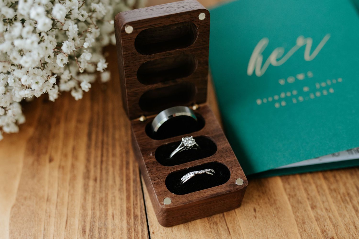Wedding rings displayed in wooden box next to turquoise book and white flowers on wooden surface.