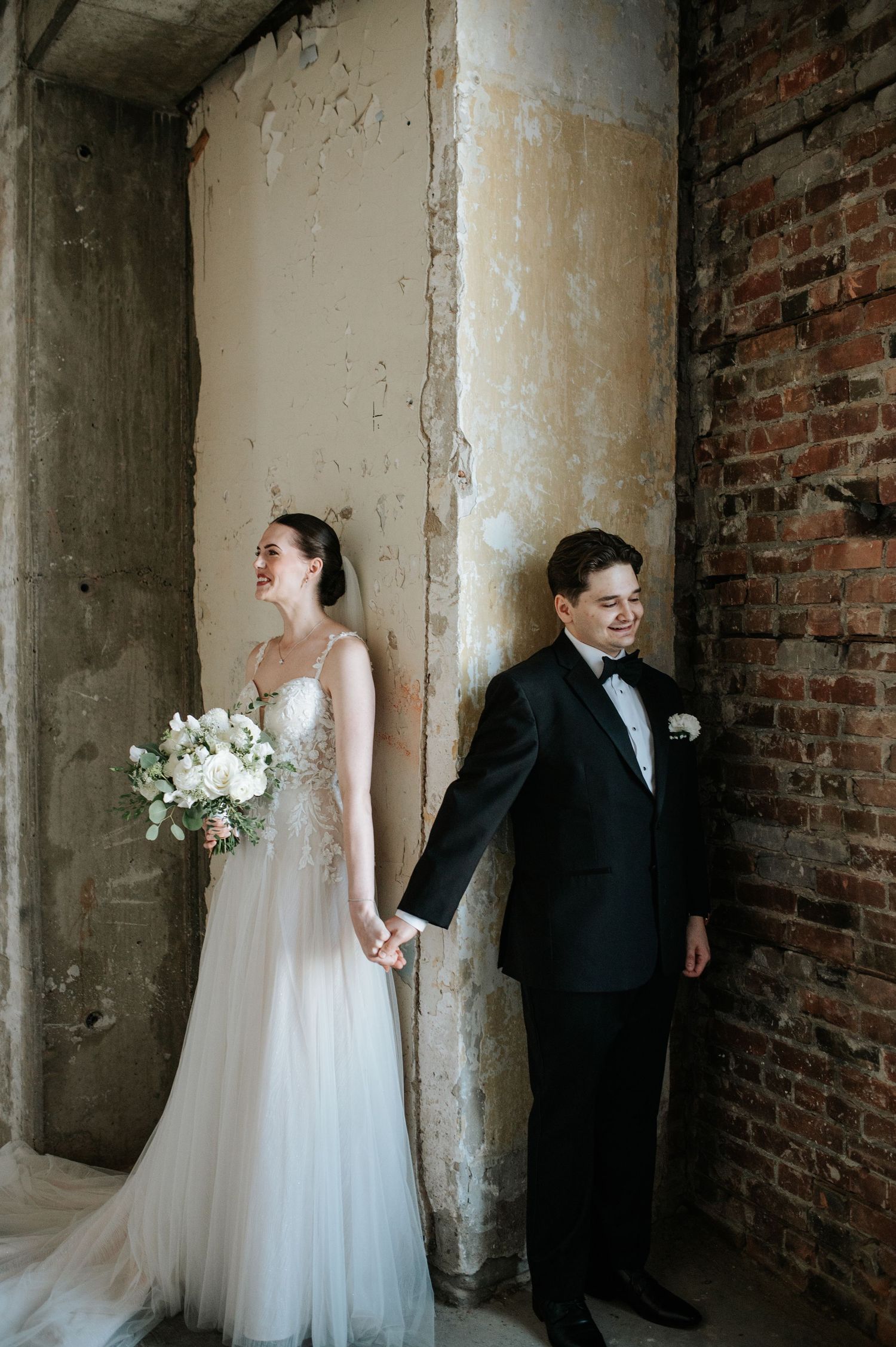 A bride in a flowing white gown and groom in classic tuxedo hold hands while standing against rustic brick walls.