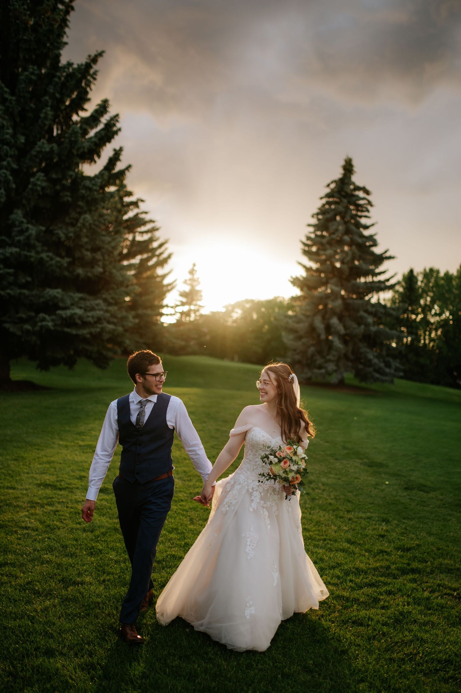 A couple walks hand in hand across a lush green lawn during golden hour with evergreen trees silhouetted against the sunset.