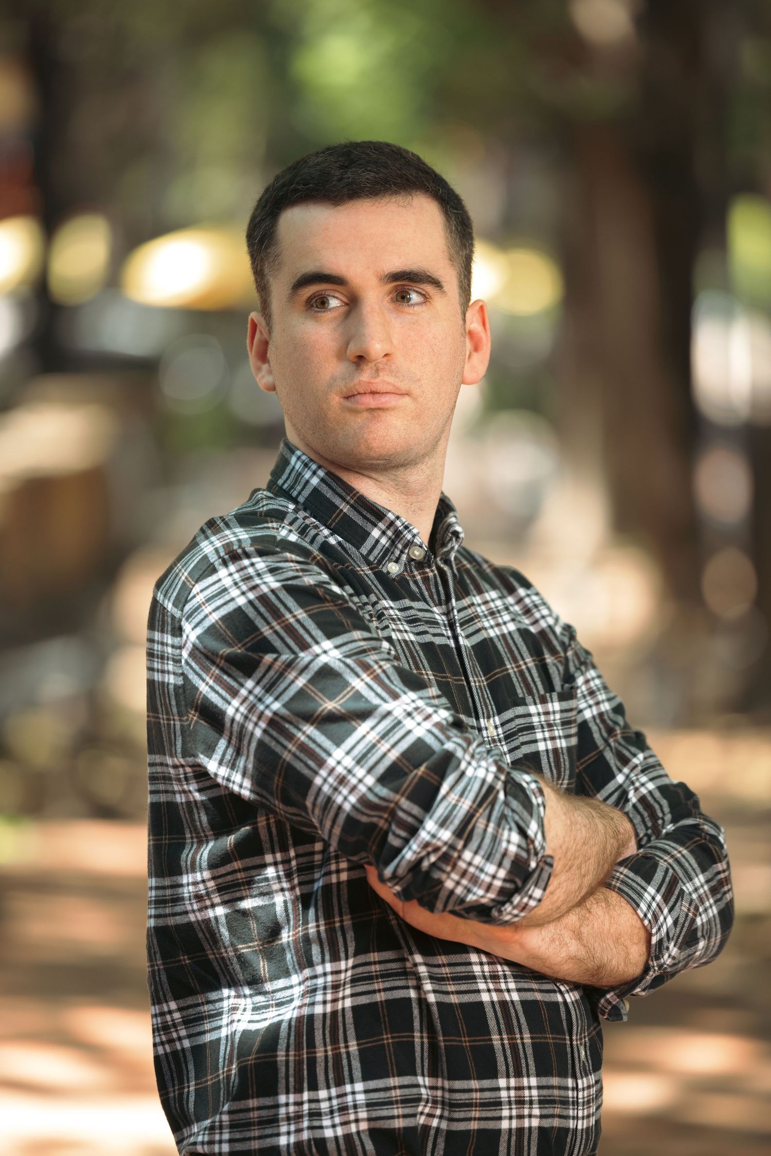 Person in black and white plaid flannel shirt poses with arms crossed in outdoor setting with blurred trees.