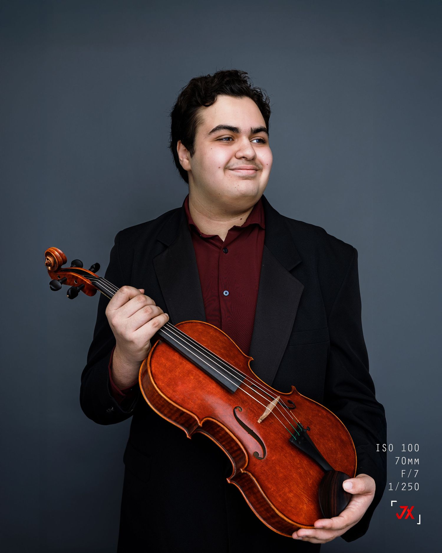 Young male violinist in black suit holding violin, smiling confidently against gray studio background.