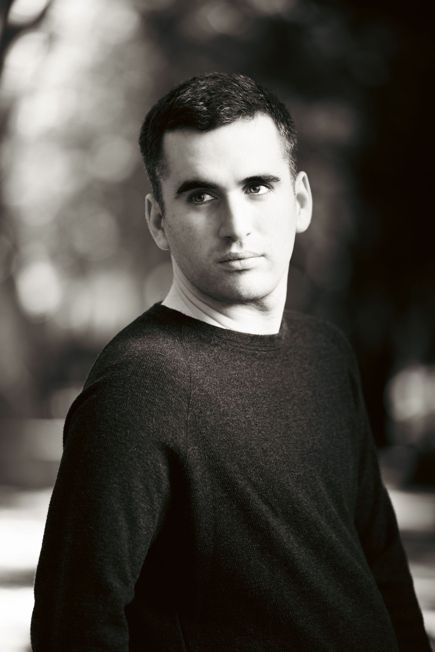 A black and white portrait of a person wearing a dark sweater looking over their shoulder against a blurred background.