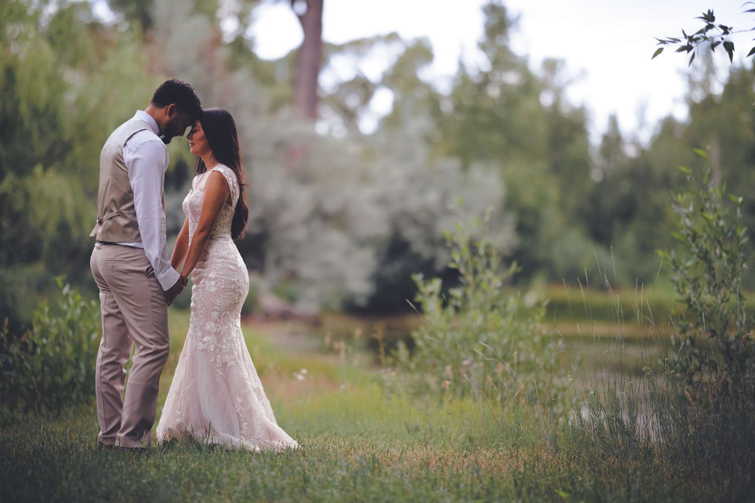 A bride and groom hold hands and stand close together in a peaceful, green outdoor setting near a pond.
