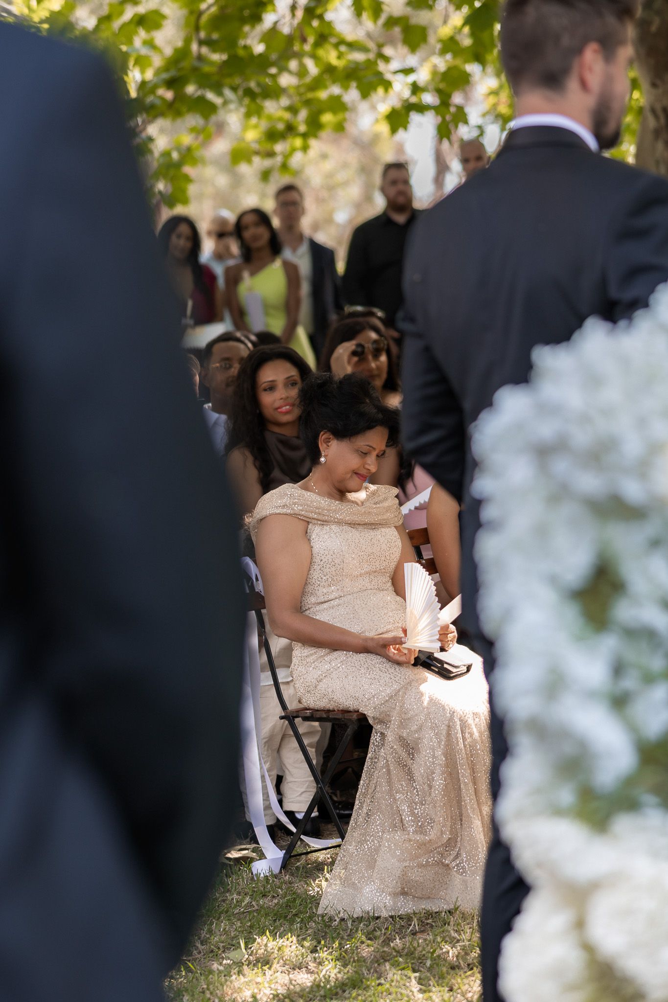 A woman in a gold lace gown sits emotionally at an outdoor wedding ceremony surrounded by guests.